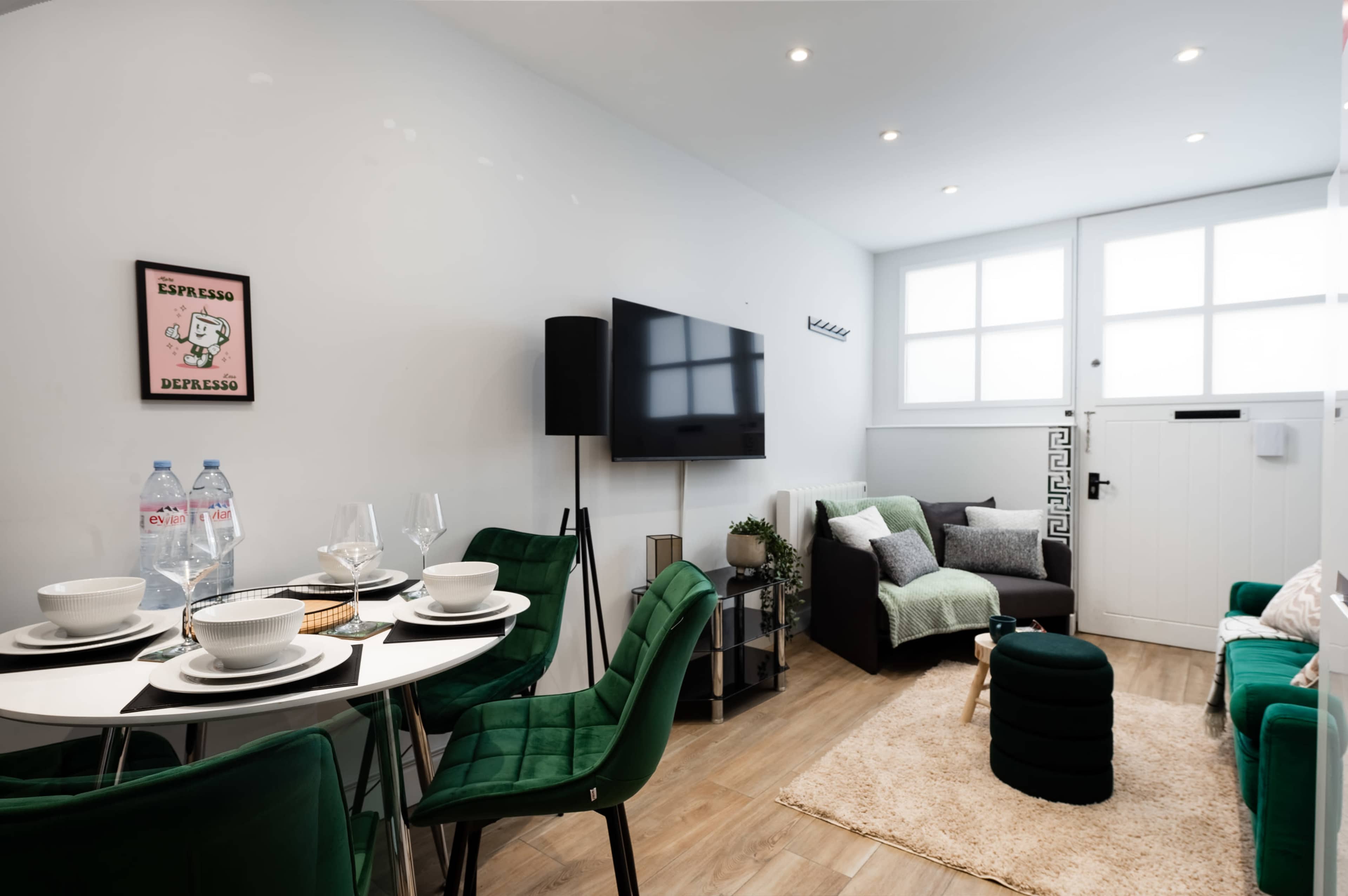 The image shows a modern, minimalistic living space featuring a dining area with a round table set for four and a cozy seating area with a television and green accents.