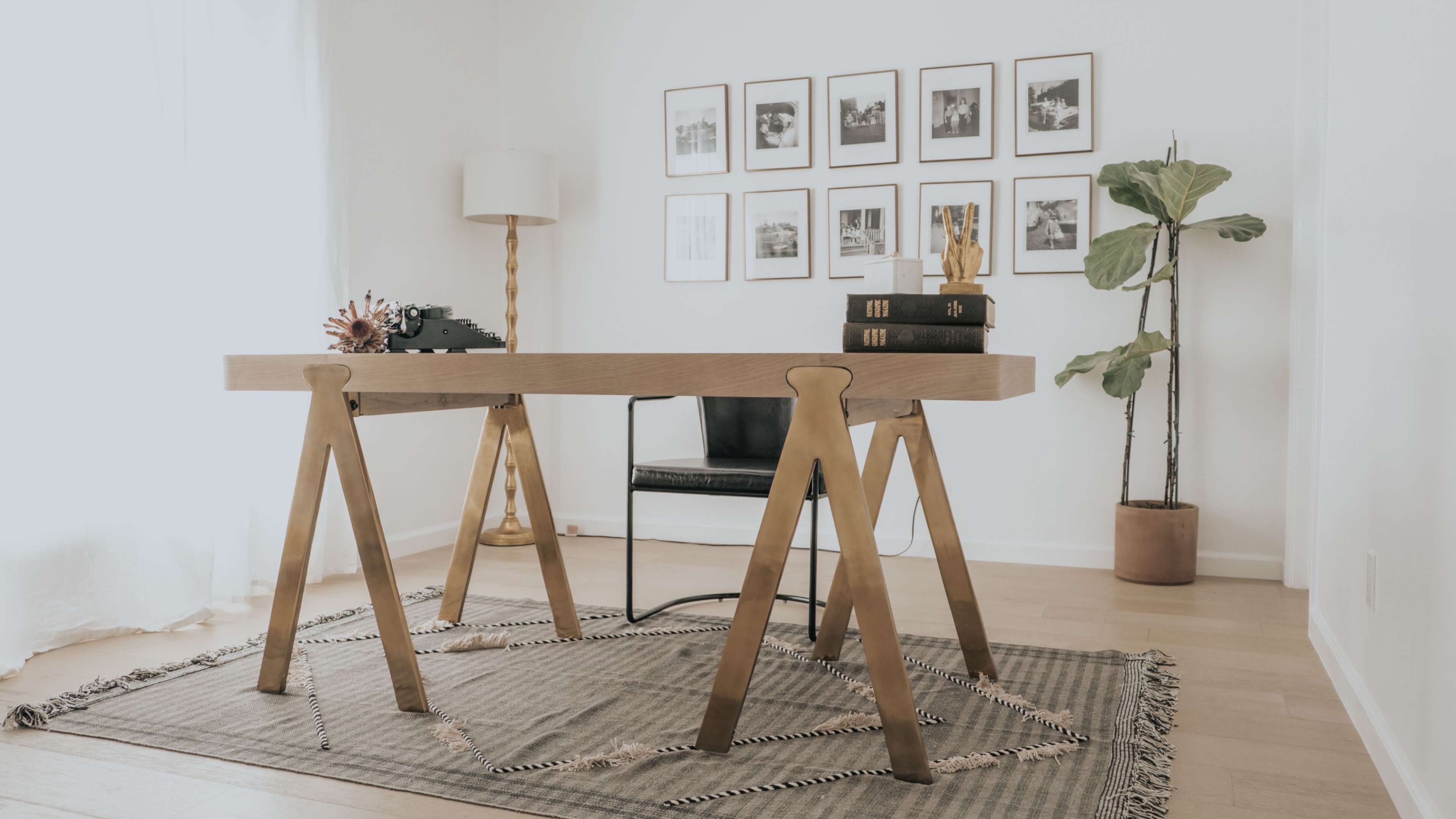 A wooden desk with sawhorse legs is positioned on a patterned rug, accompanied by a chair, plants, and framed photos on the wall.