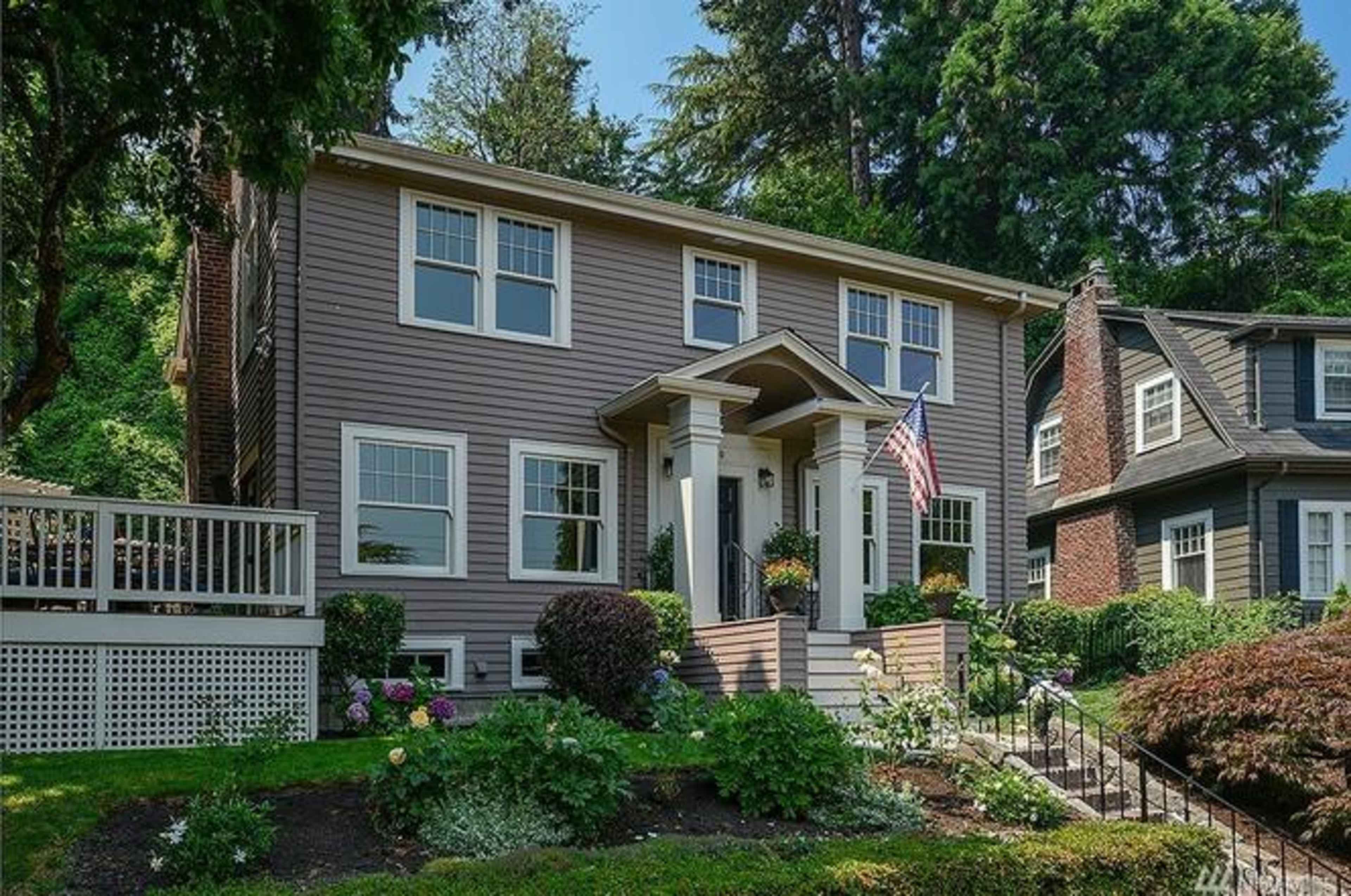 A two-story house with a light gray facade, featuring a front porch and an American flag, surrounded by landscaped greenery and flowers.