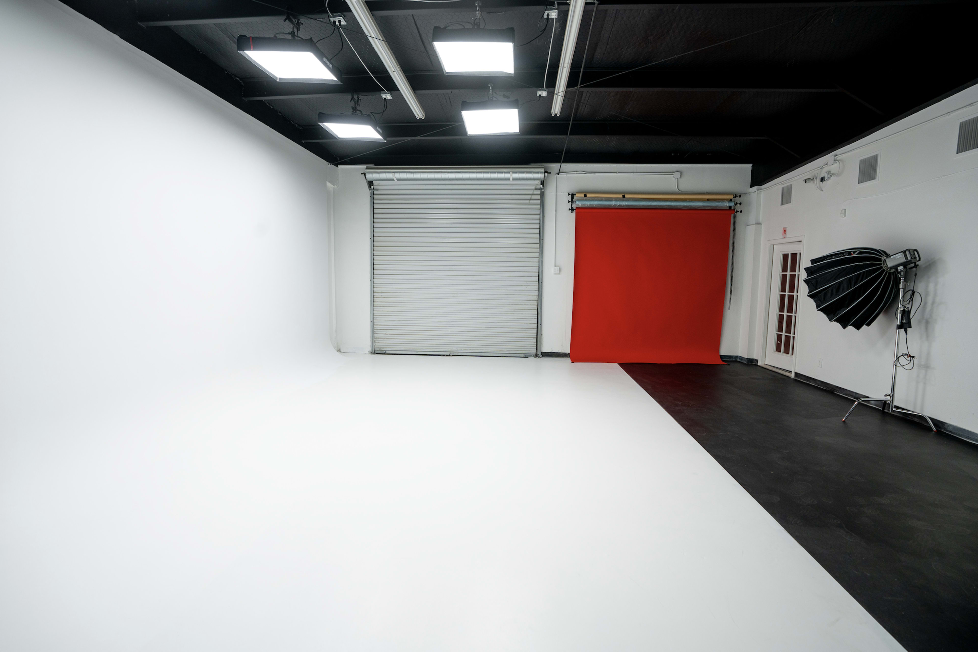 The image shows a photography studio with a white backdrop on one side and a red backdrop on the other, along with a folding umbrella light setup.