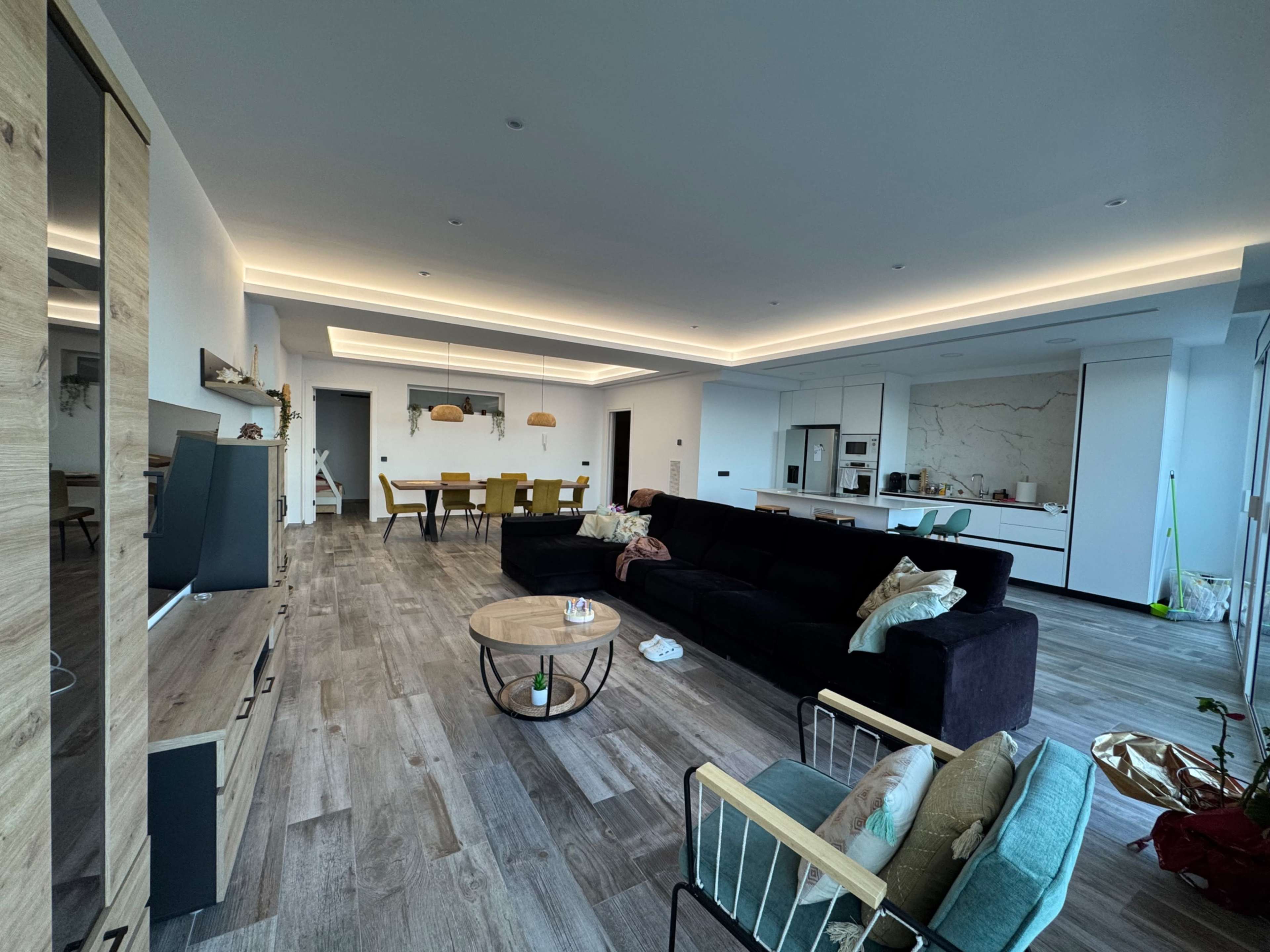 The image shows a modern living room with a black sofa, wooden flooring, and a dining area in the background.