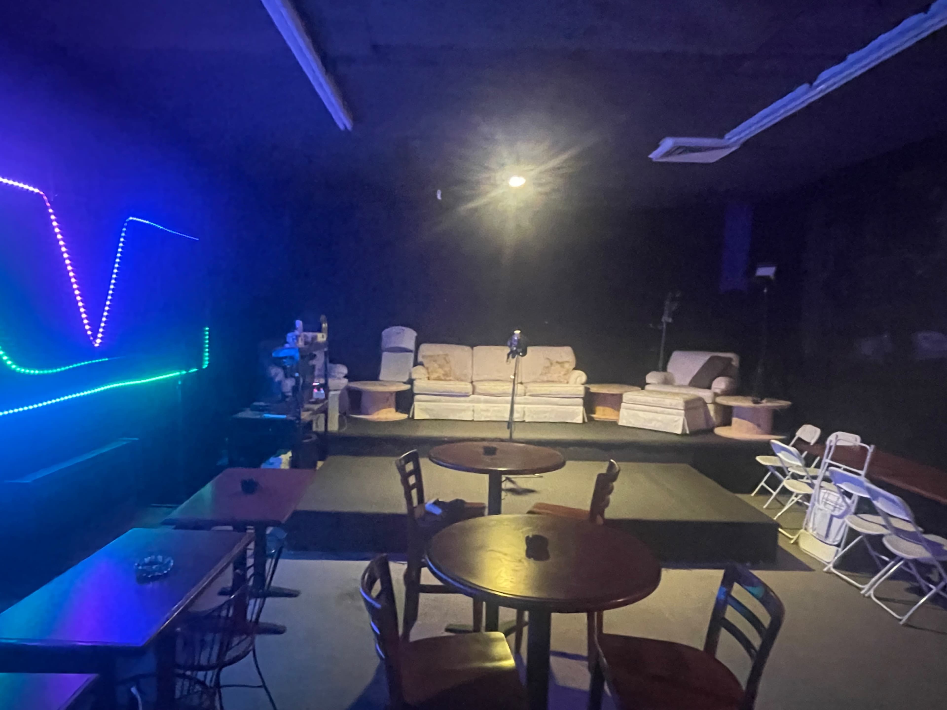The image shows an empty performance venue with round tables and a small stage featuring a microphone, surrounded by couches and chairs.