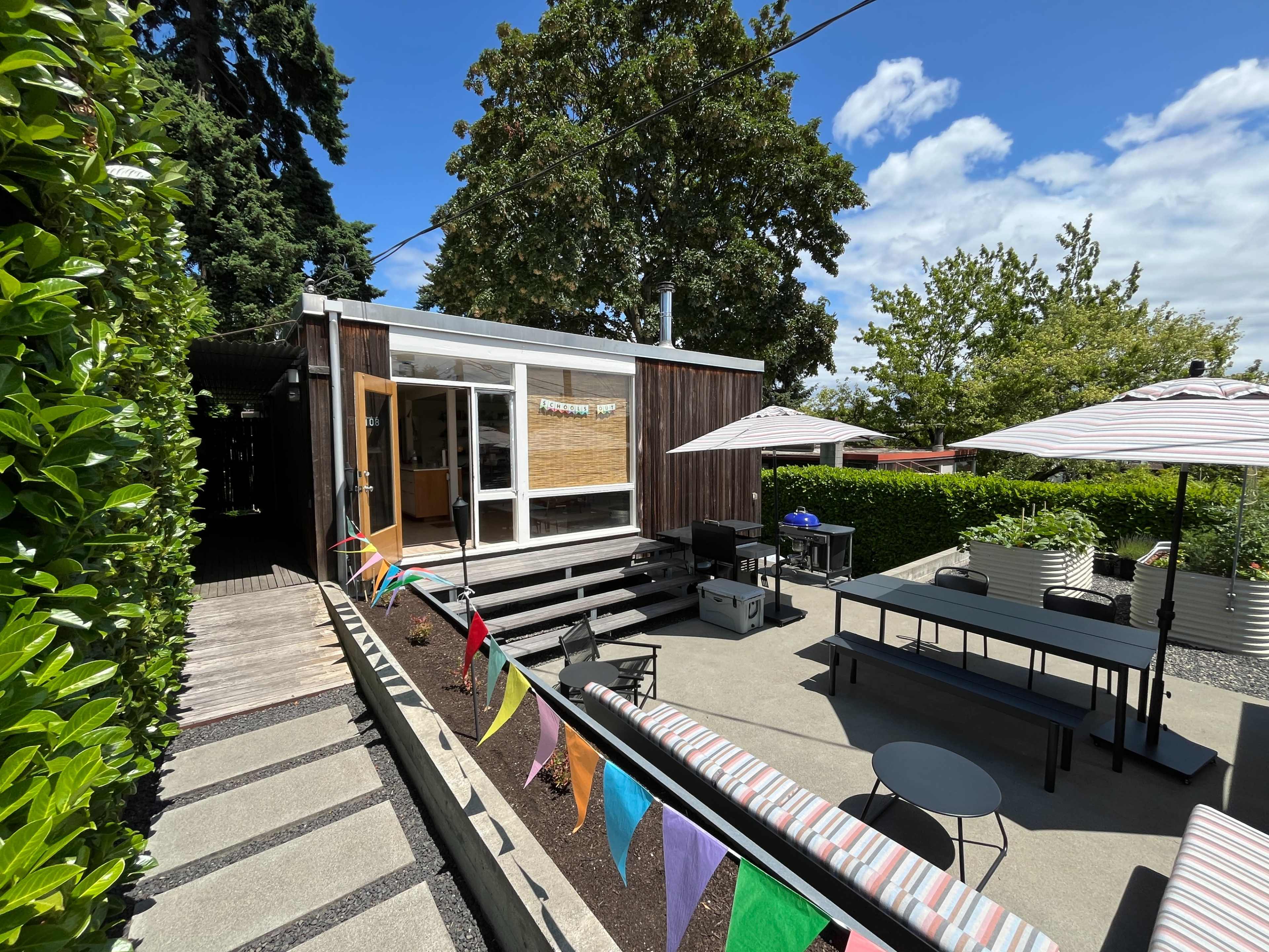 The image shows a modern, one-story house with large windows, surrounded by greenery and patio furniture, featuring colorful bunting strung across a concrete walking path.