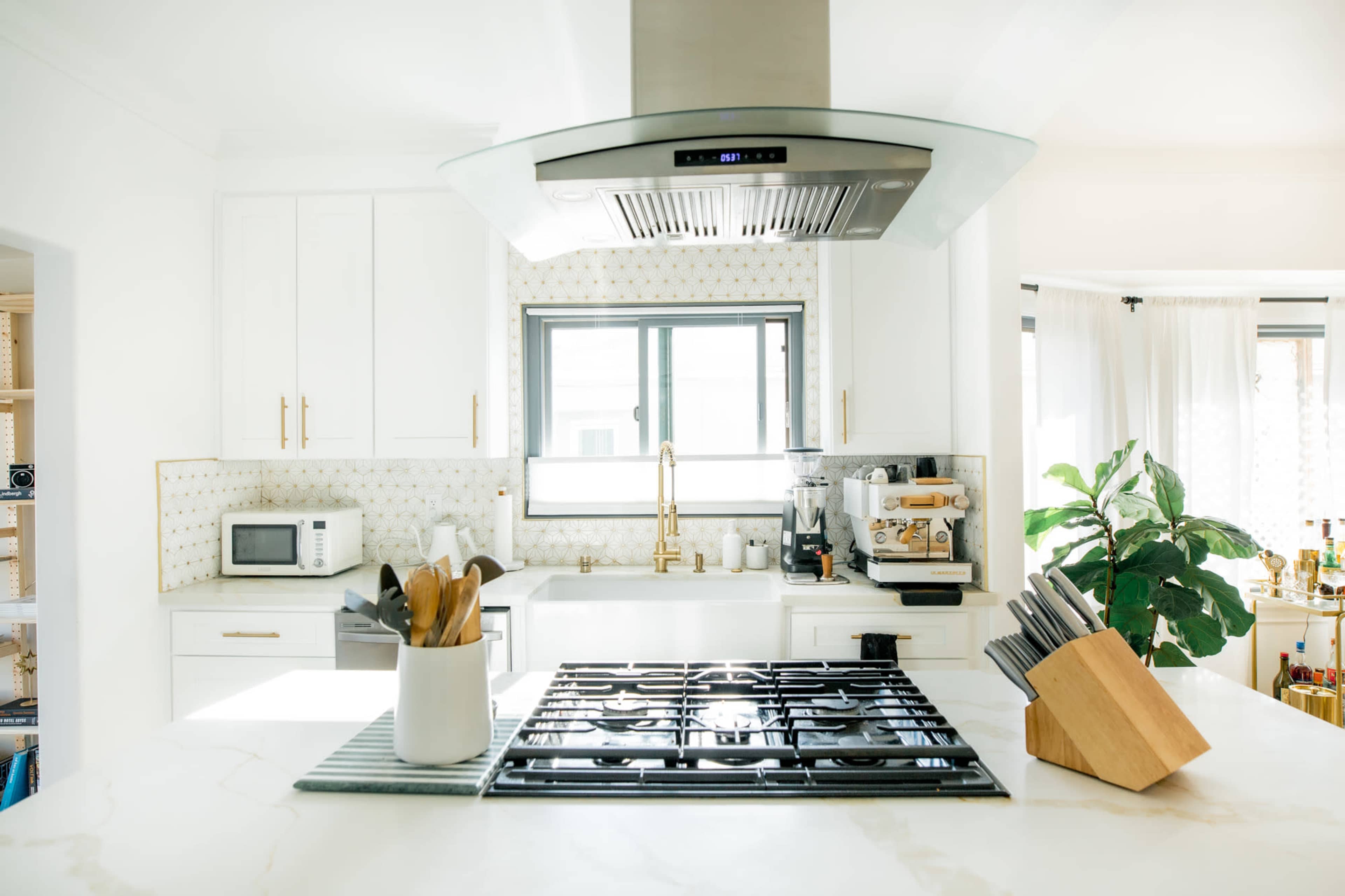 The image shows a modern kitchen featuring a gas stovetop, a range hood above, and various kitchen appliances, including a microwave and coffee machine, with a window providing natural light.