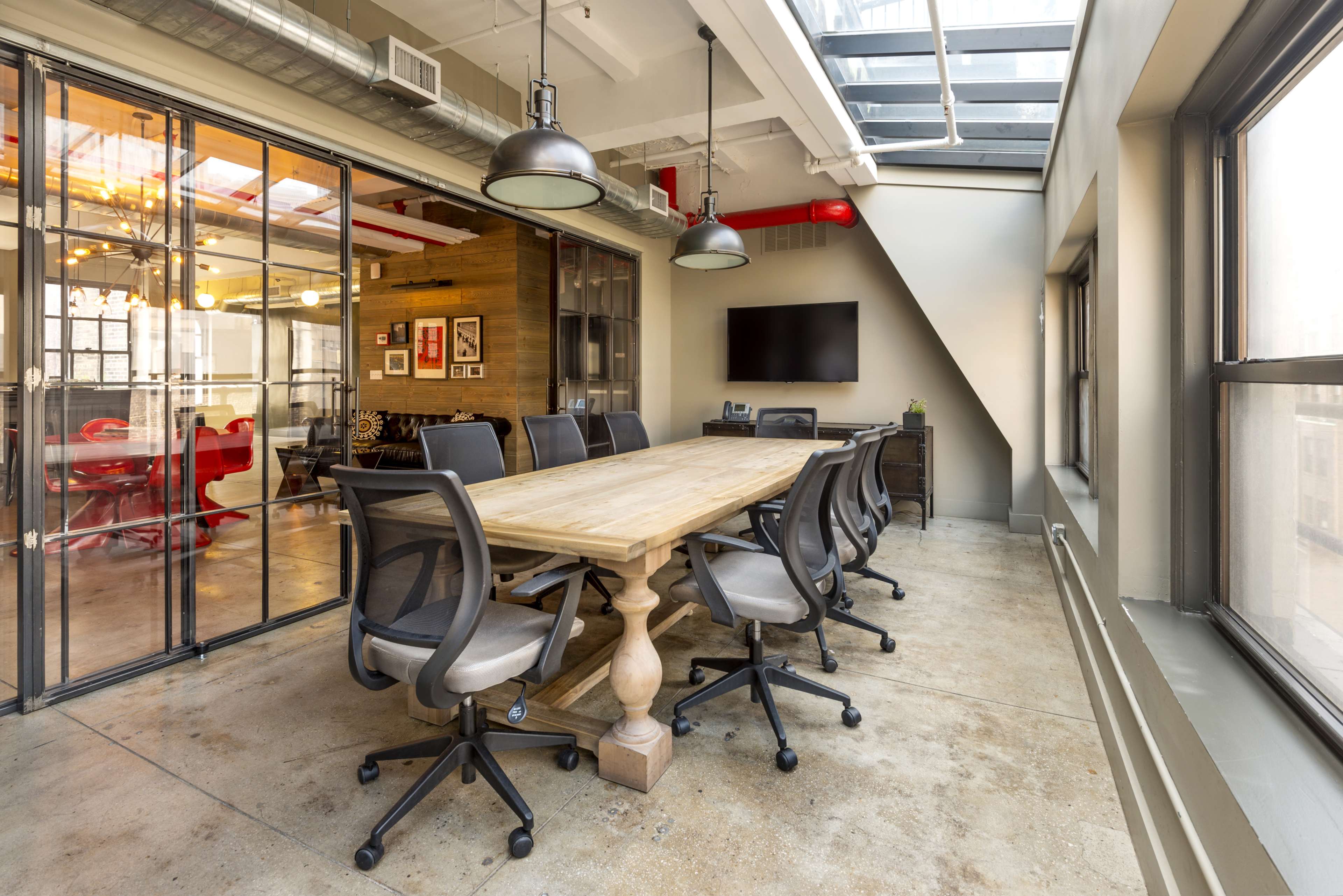 A modern conference room features a large wooden table surrounded by ergonomic chairs, with glass walls and a wall-mounted display.