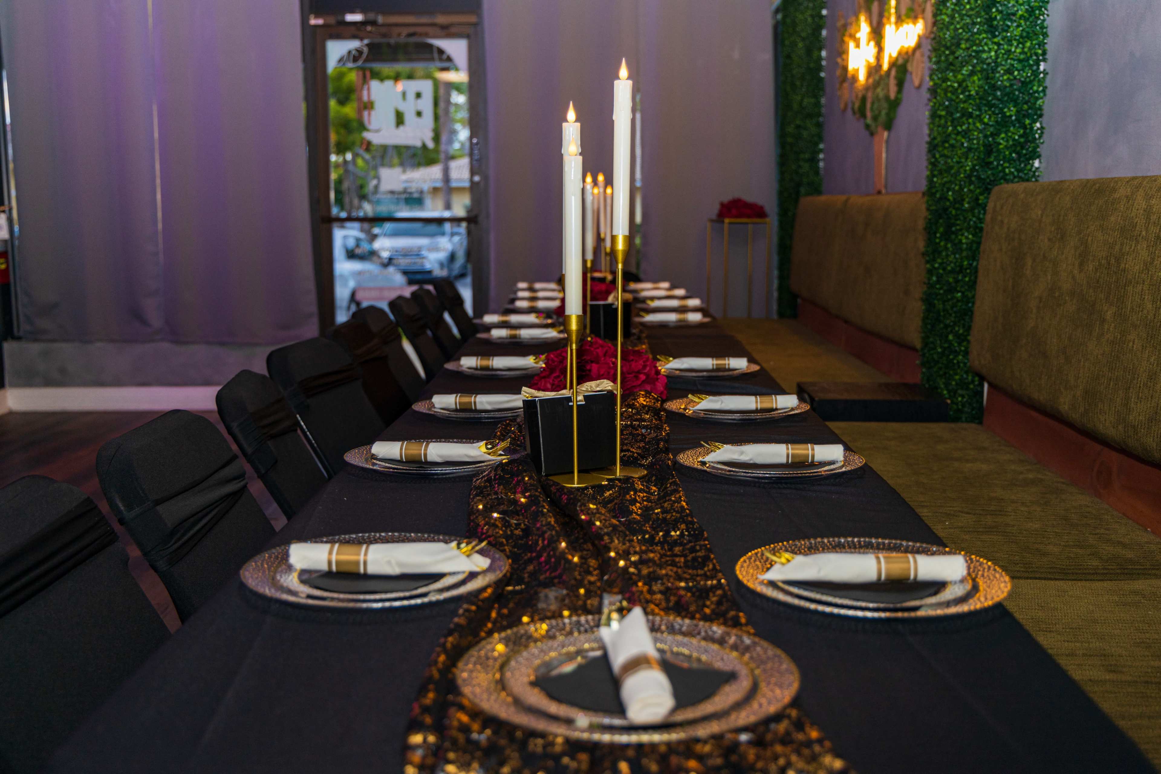 A long dining table is set with elegant black and gold decorations, featuring candles and plates arranged along a shimmering table runner.