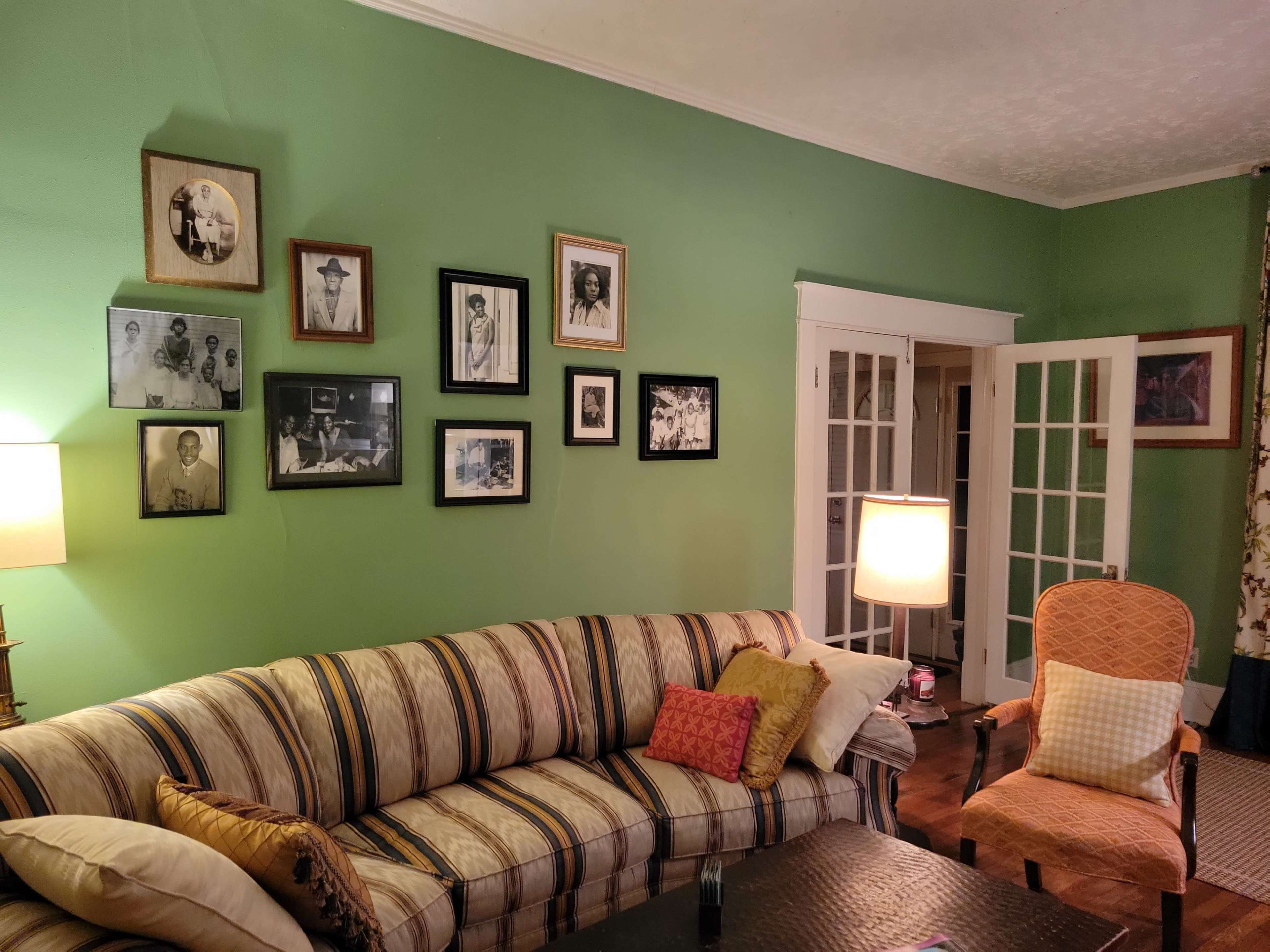 A green living room features a striped sofa, a lamp, and a wall decorated with framed black and white photographs.