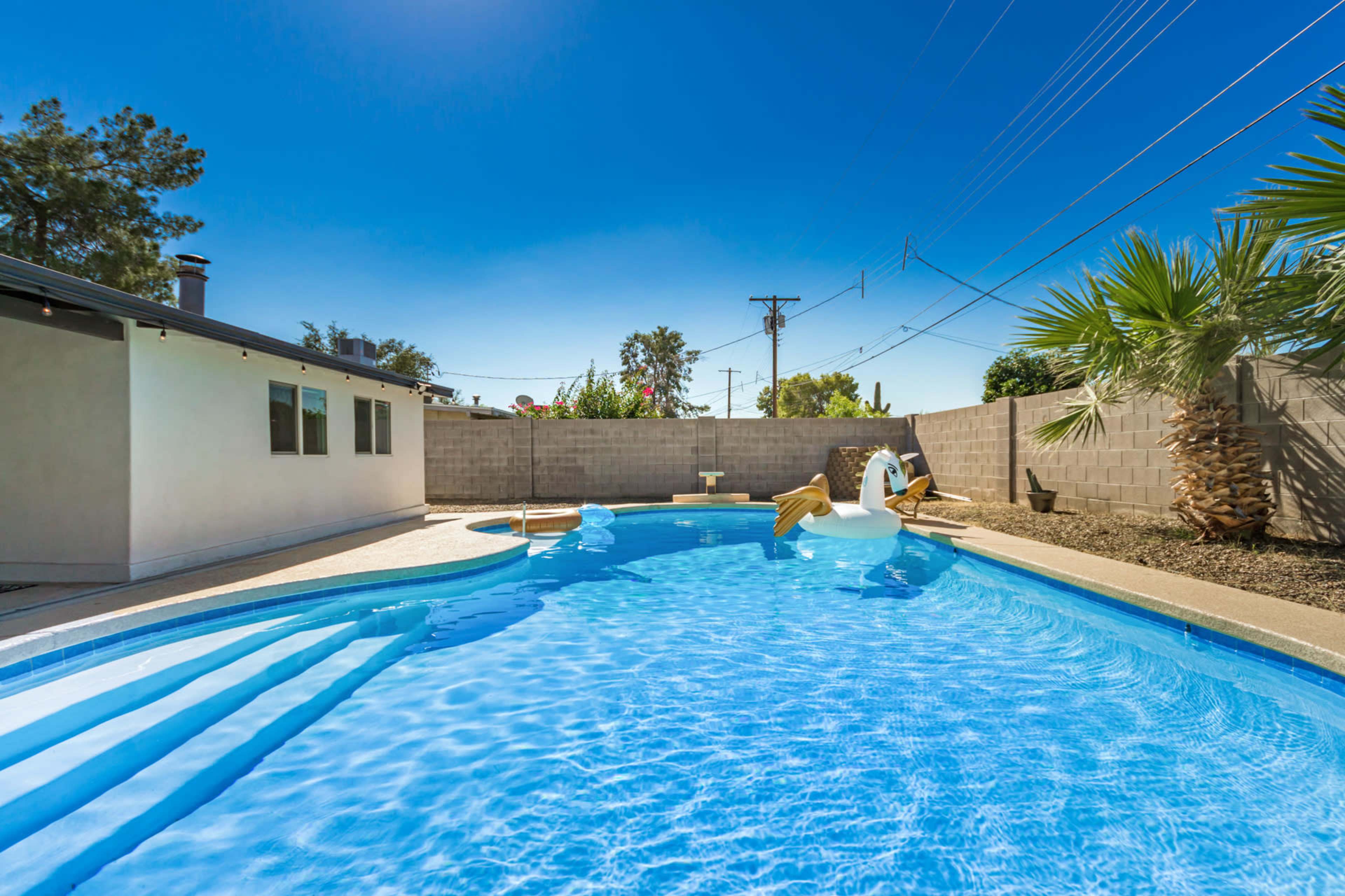 A clear swimming pool with a slide and a large inflatable swan is situated in a backyard with palm trees and a brick wall.