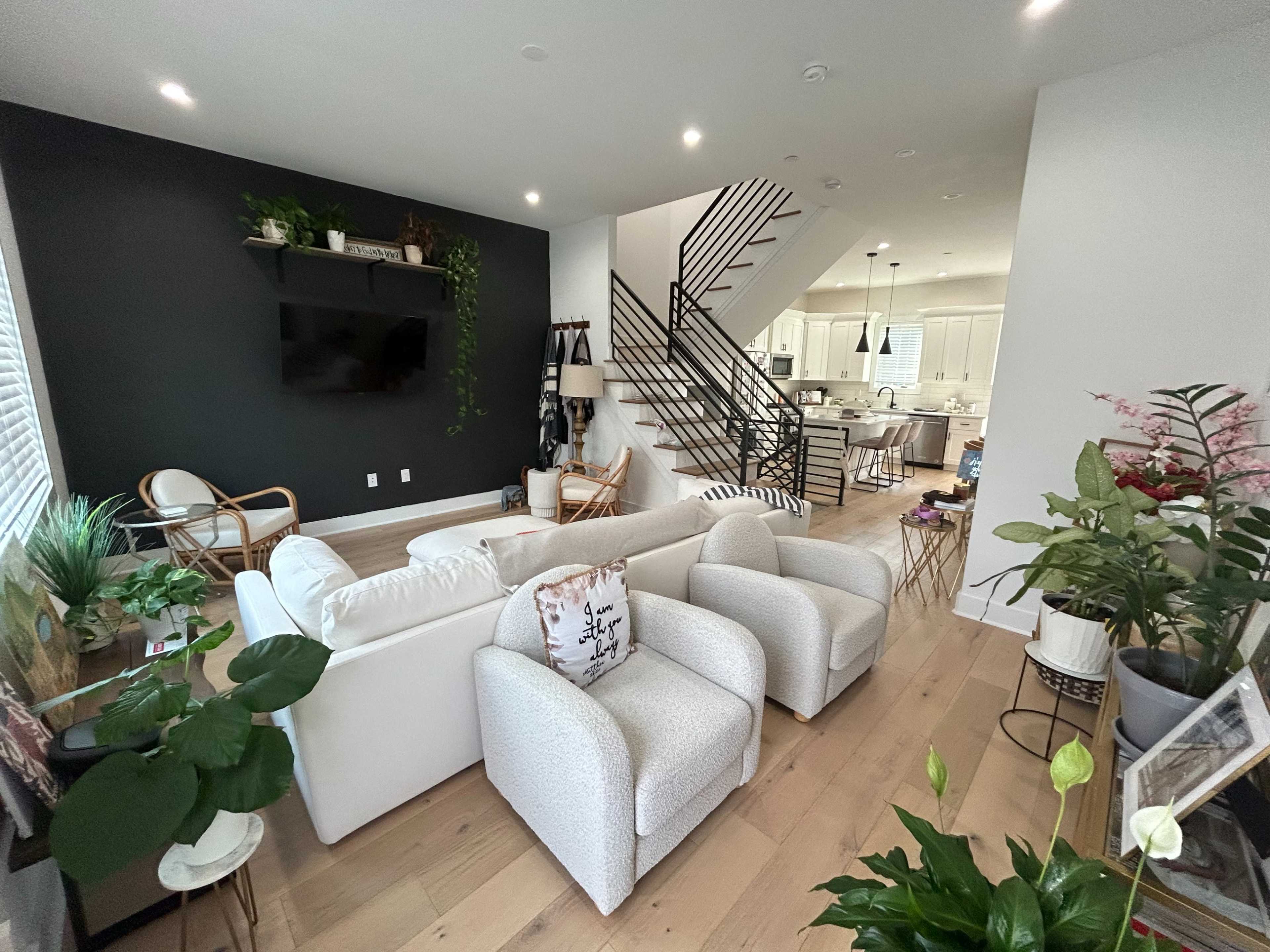 The image shows a modern living space with a white sofa, two armchairs, a staircase leading to an upper level, and a kitchen in the background.