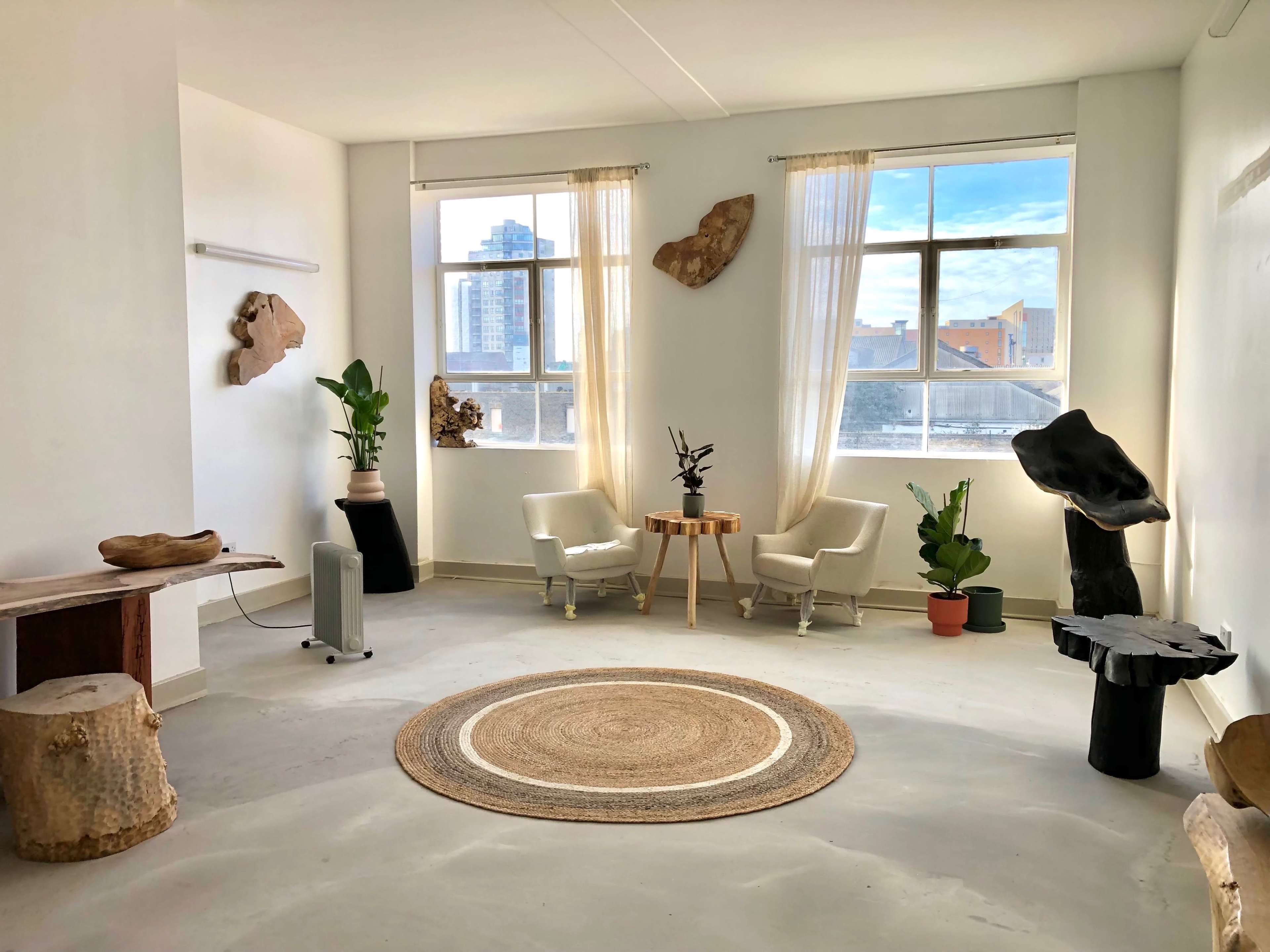A minimalist room with two chairs and a round rug, featuring wooden sculptures on the walls and plants in the corners.