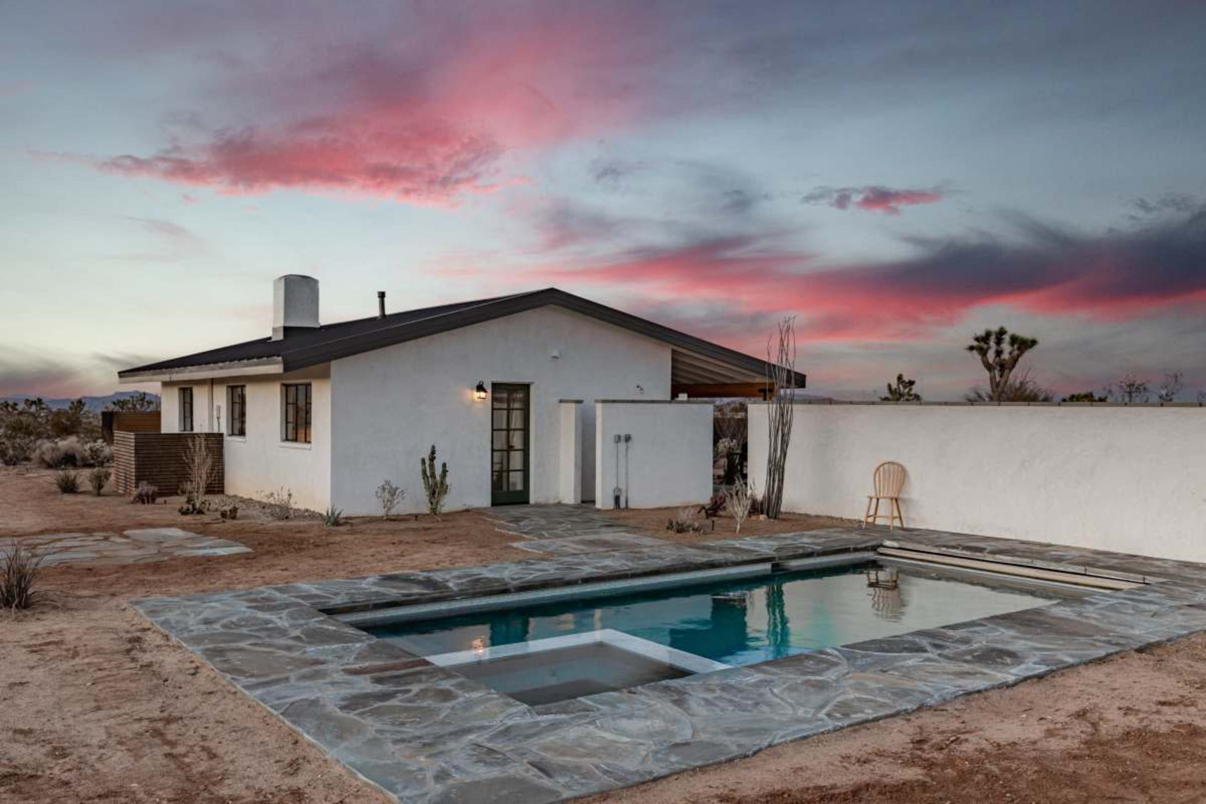 A modern single-story house with a stone patio and swimming pool is set against a colorful sky at dusk.