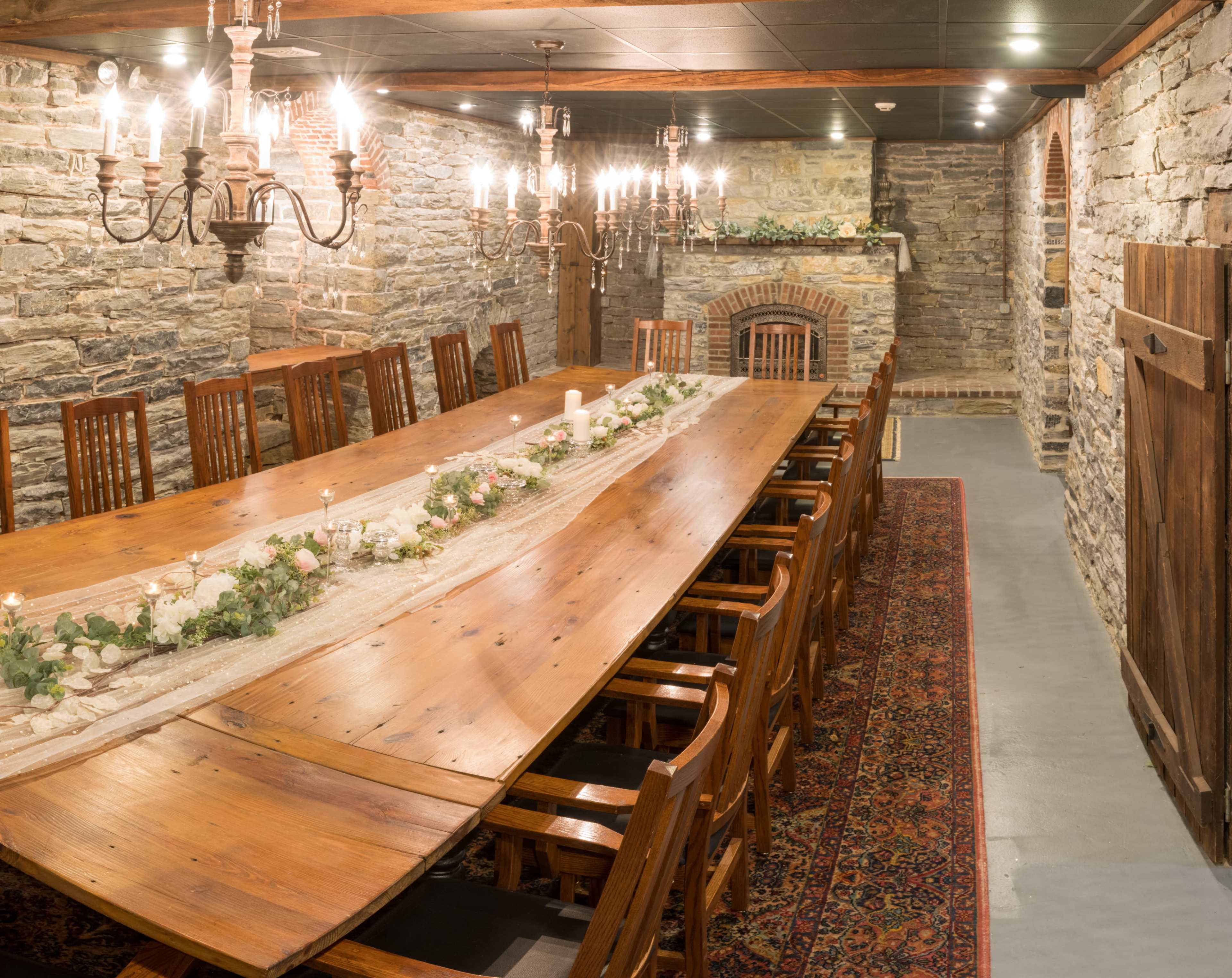 The image shows a long wooden dining table surrounded by chairs in a rustic room with stone walls and multiple chandeliers.