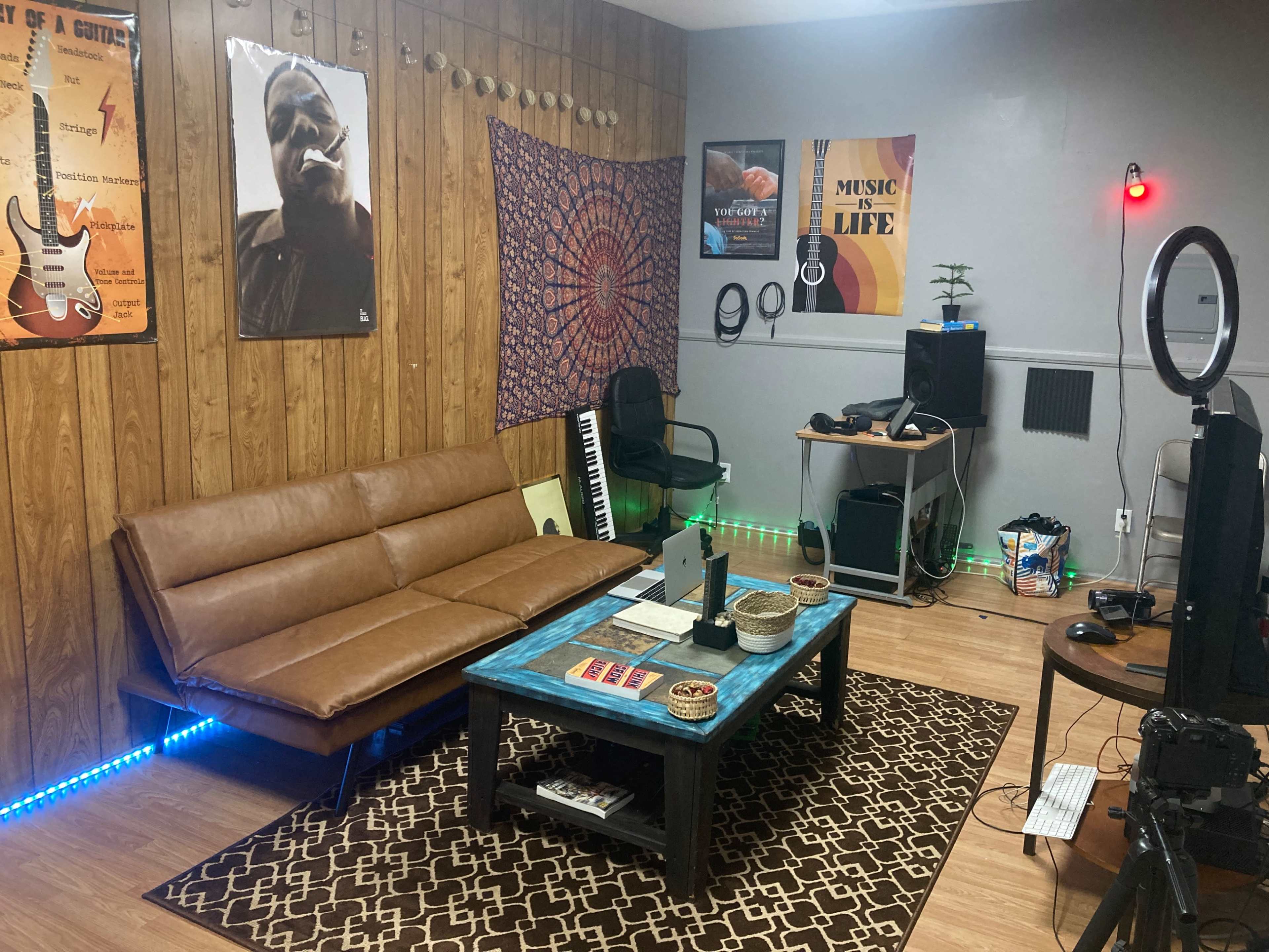 The image shows a music-themed room featuring a brown sofa, a blue coffee table, posters on the walls, and various musical instruments and equipment.