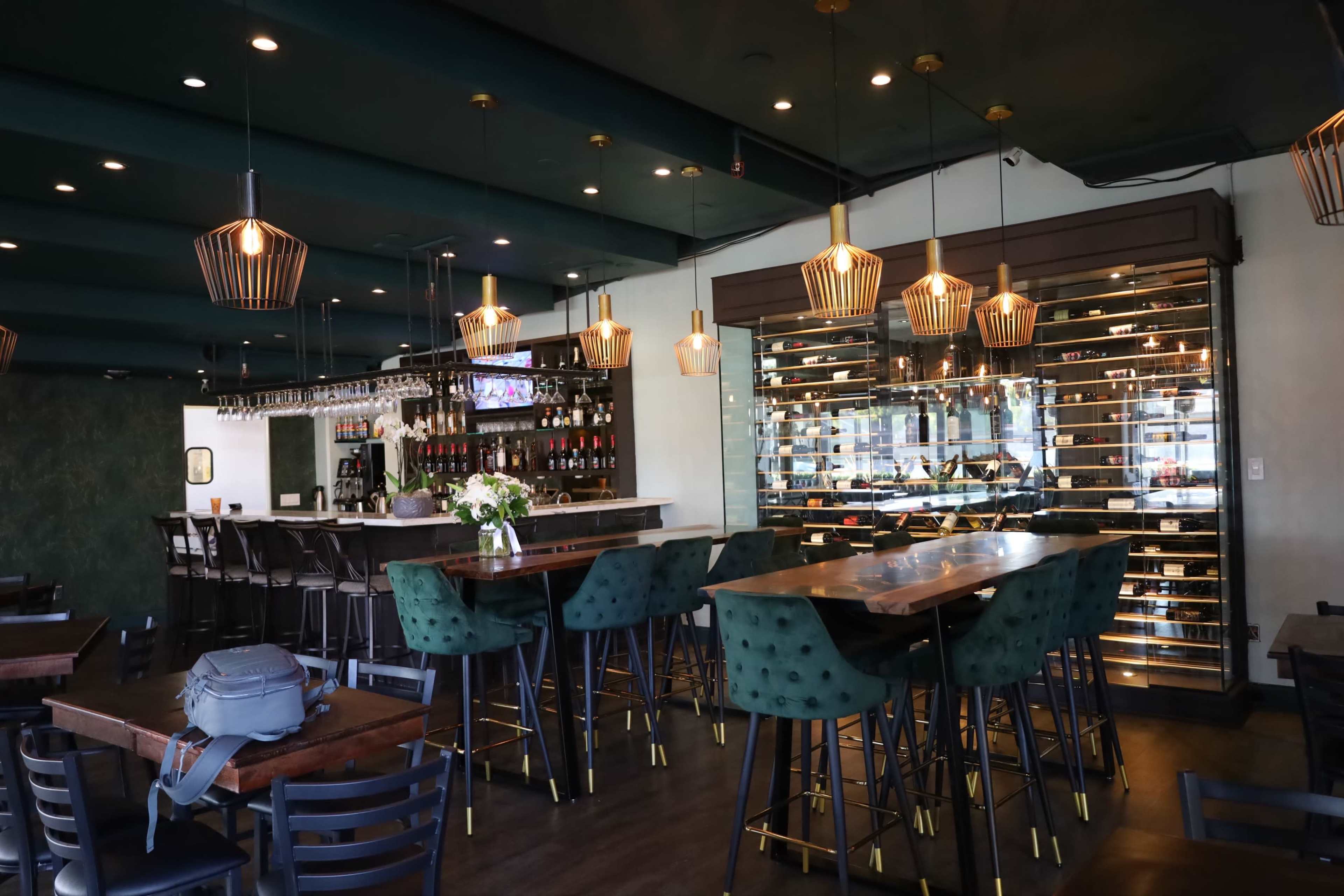 The image shows a modern restaurant interior with green and gold decor, featuring a bar area with tall stools and a wine display behind the counter.