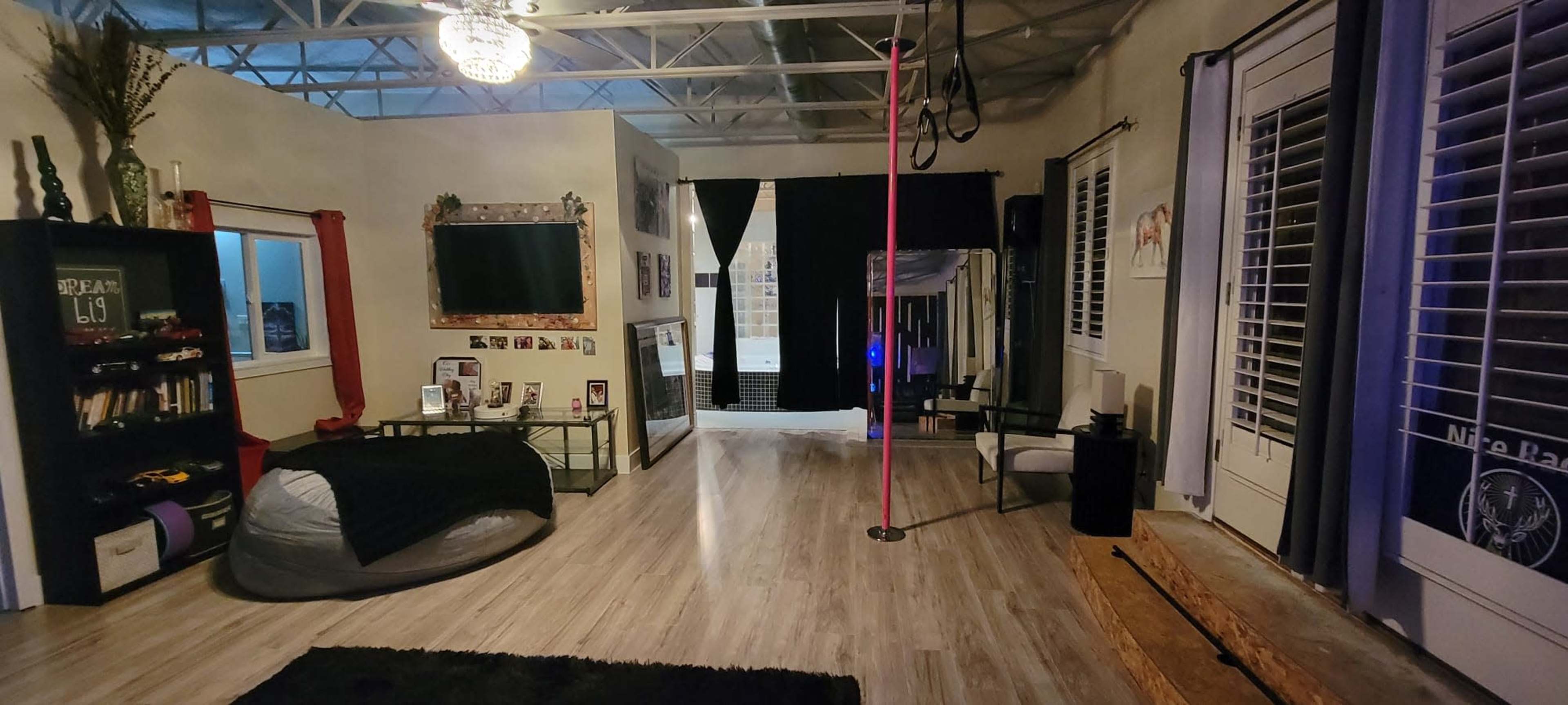 The image shows a spacious indoor area with a mirrored wall, pole in the center, and various furniture and decor items, including a large bean bag and bookshelves.