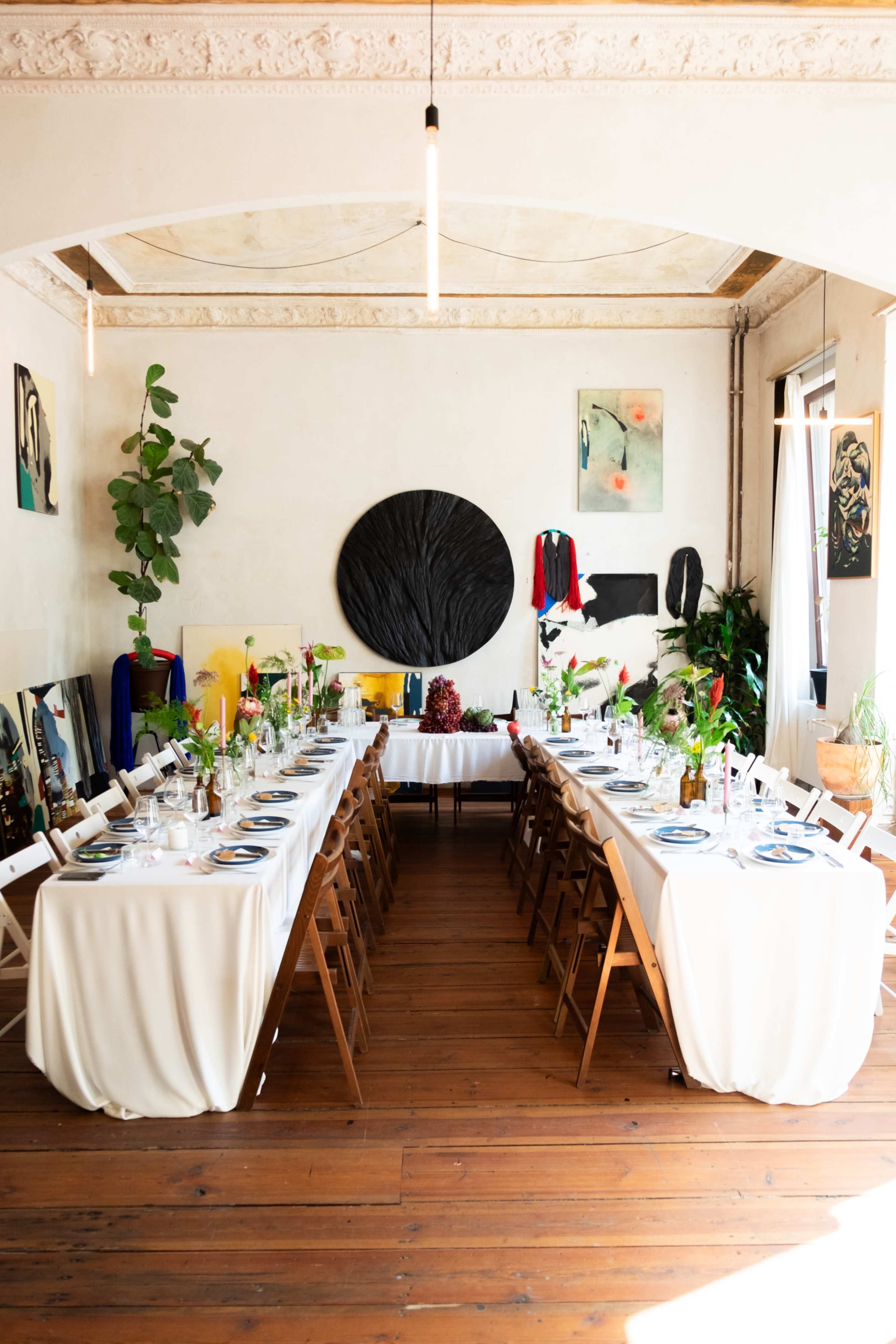 The image shows a dining room set with a long table covered in white cloth, surrounded by wooden chairs, and decorated with plants and artwork on the walls.