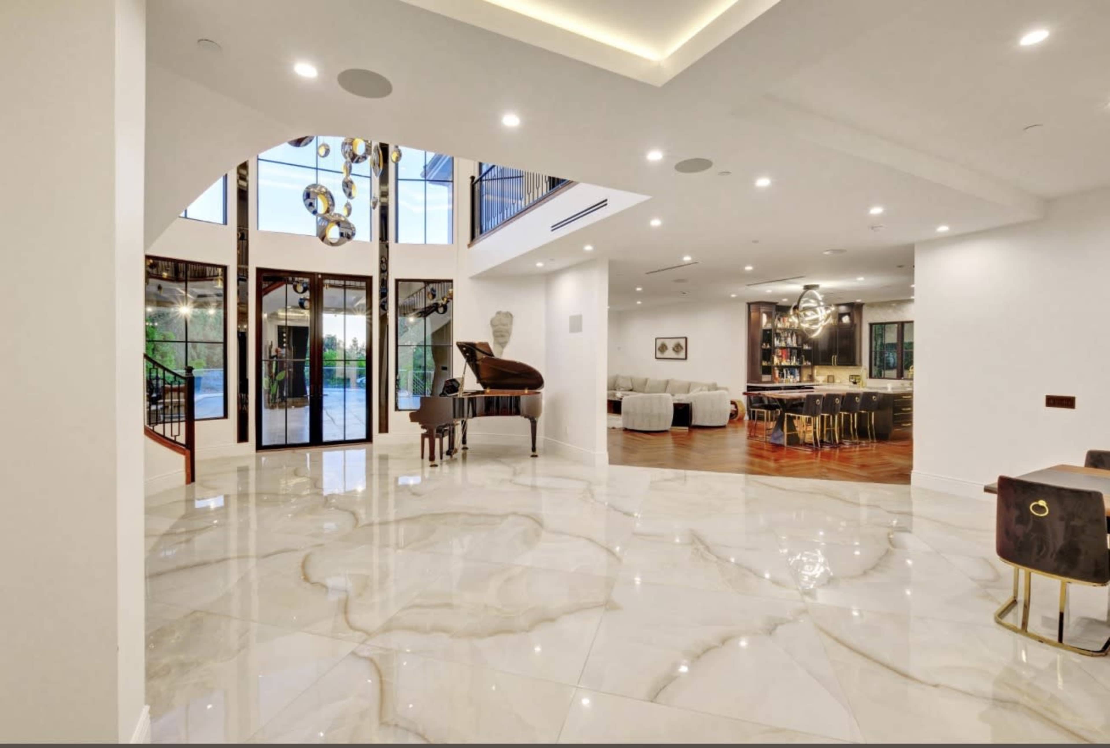The image shows a modern, open-concept living space featuring a grand piano, large windows, and polished marble floors.
