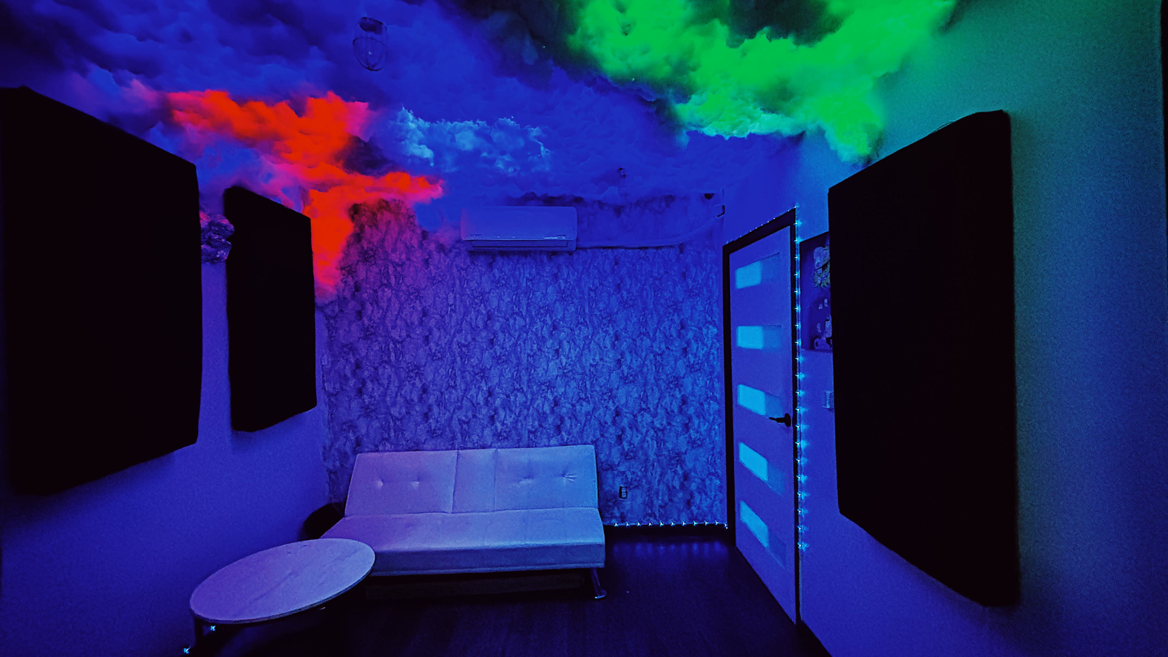 A dimly lit room features a white sofa, colorful cloud-like lighting on the ceiling, and soundproof panels on the walls.