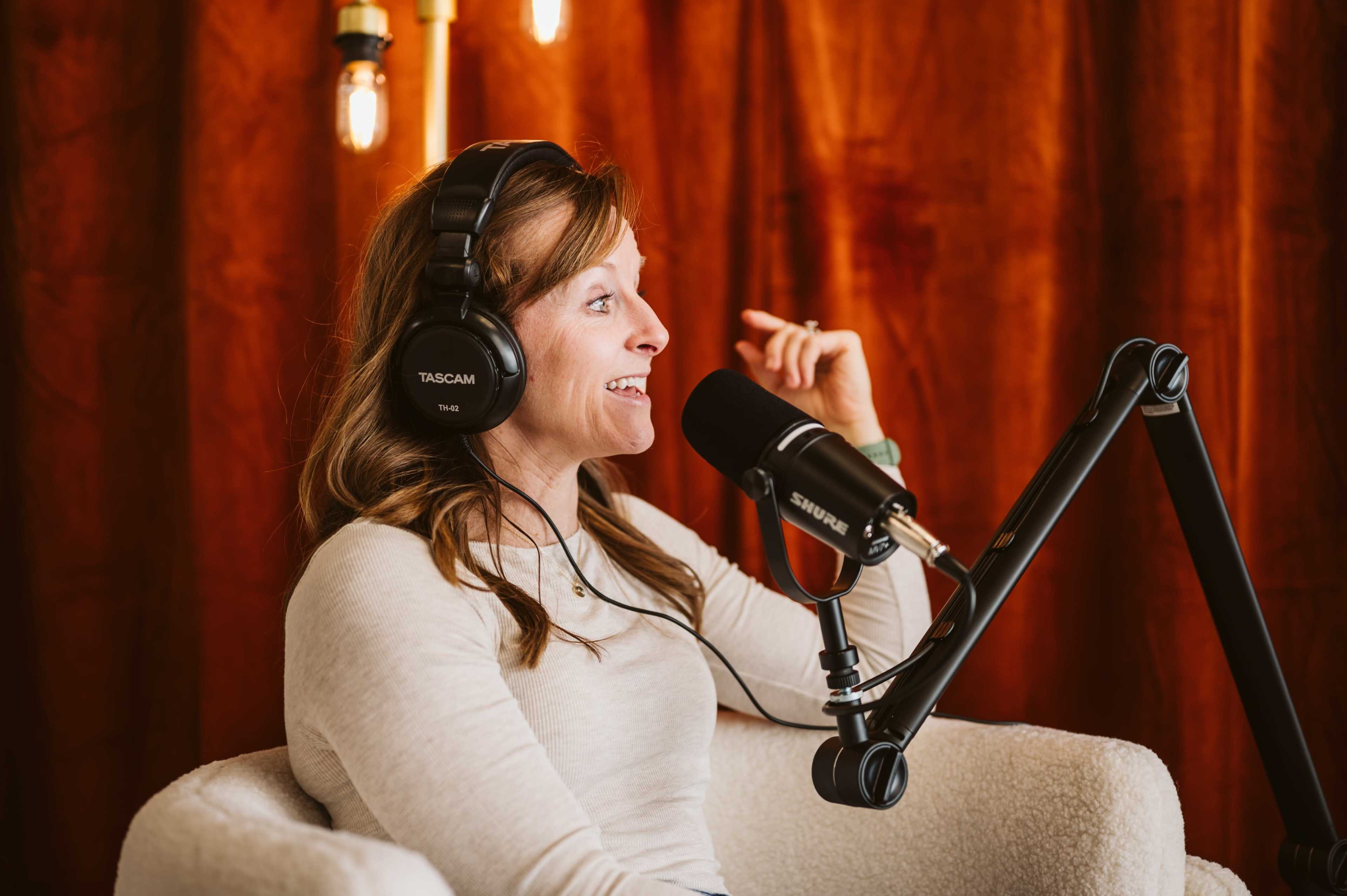 A woman with headphones smiles while speaking into a microphone in a cozy recording studio setting.