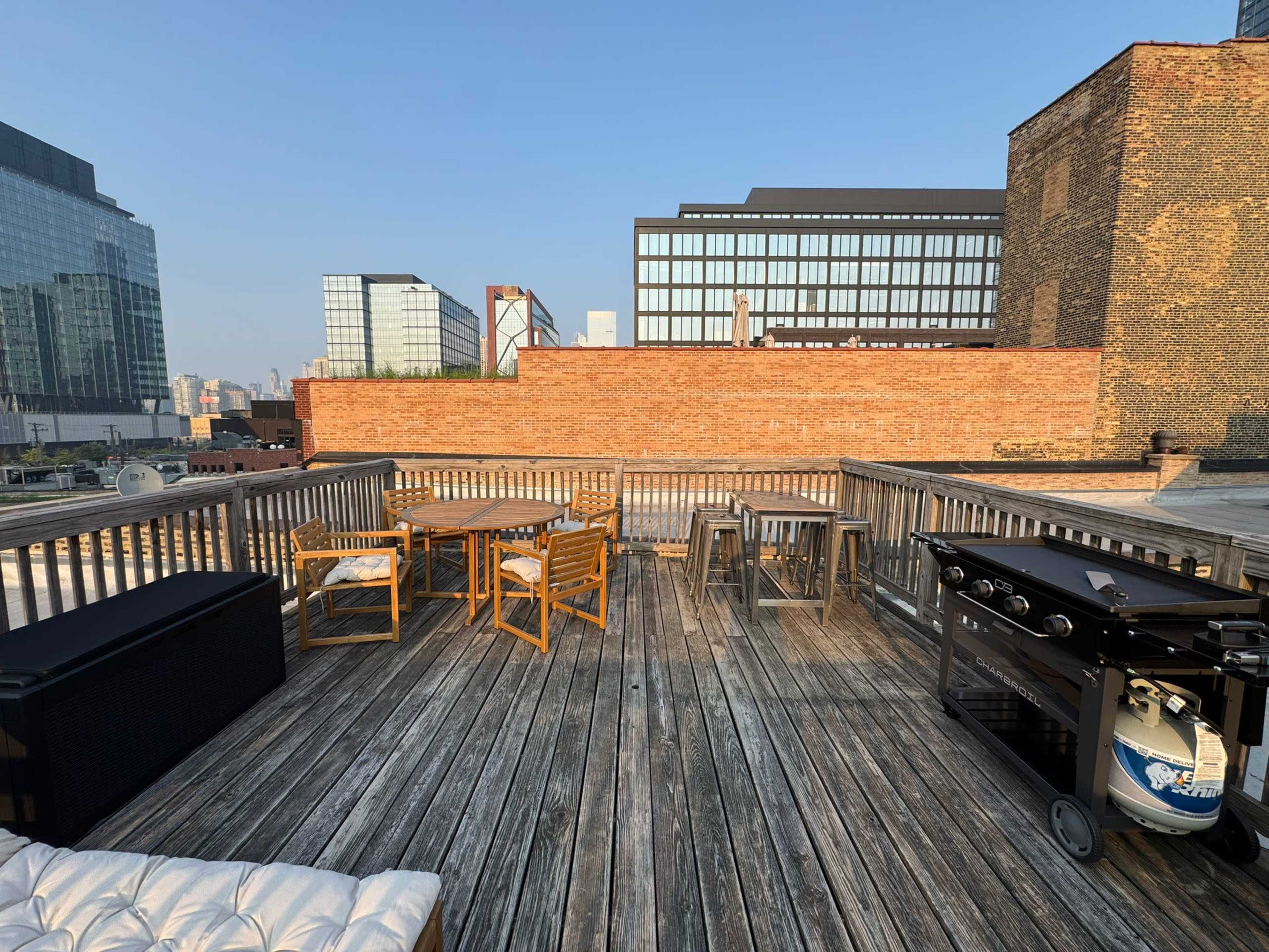 The image shows a wooden rooftop deck with several tables and chairs, along with a grill, against a backdrop of urban buildings.