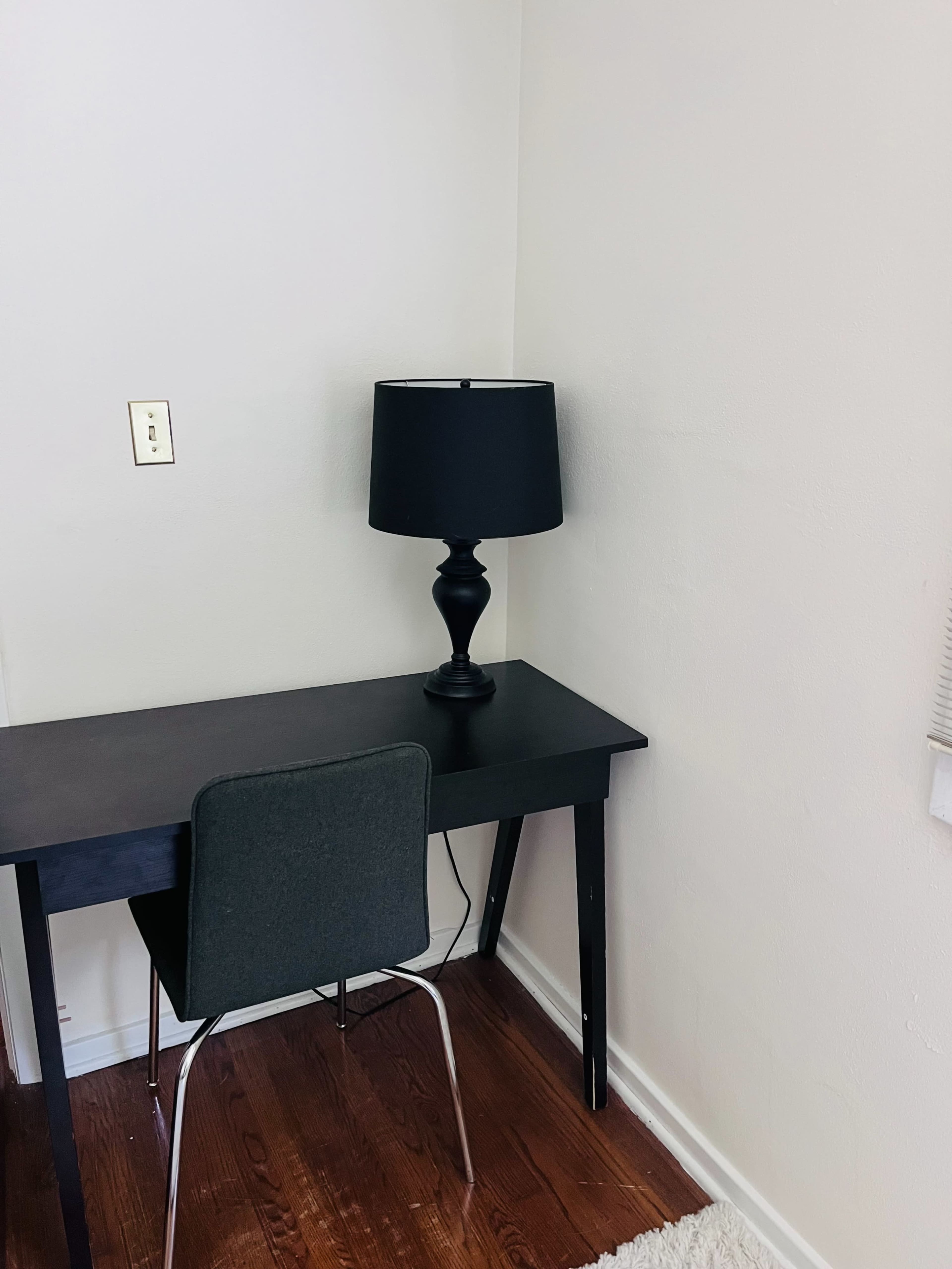 A black desk with a matching lamp and a gray chair is positioned in a corner of a room with light-colored walls and hardwood flooring.