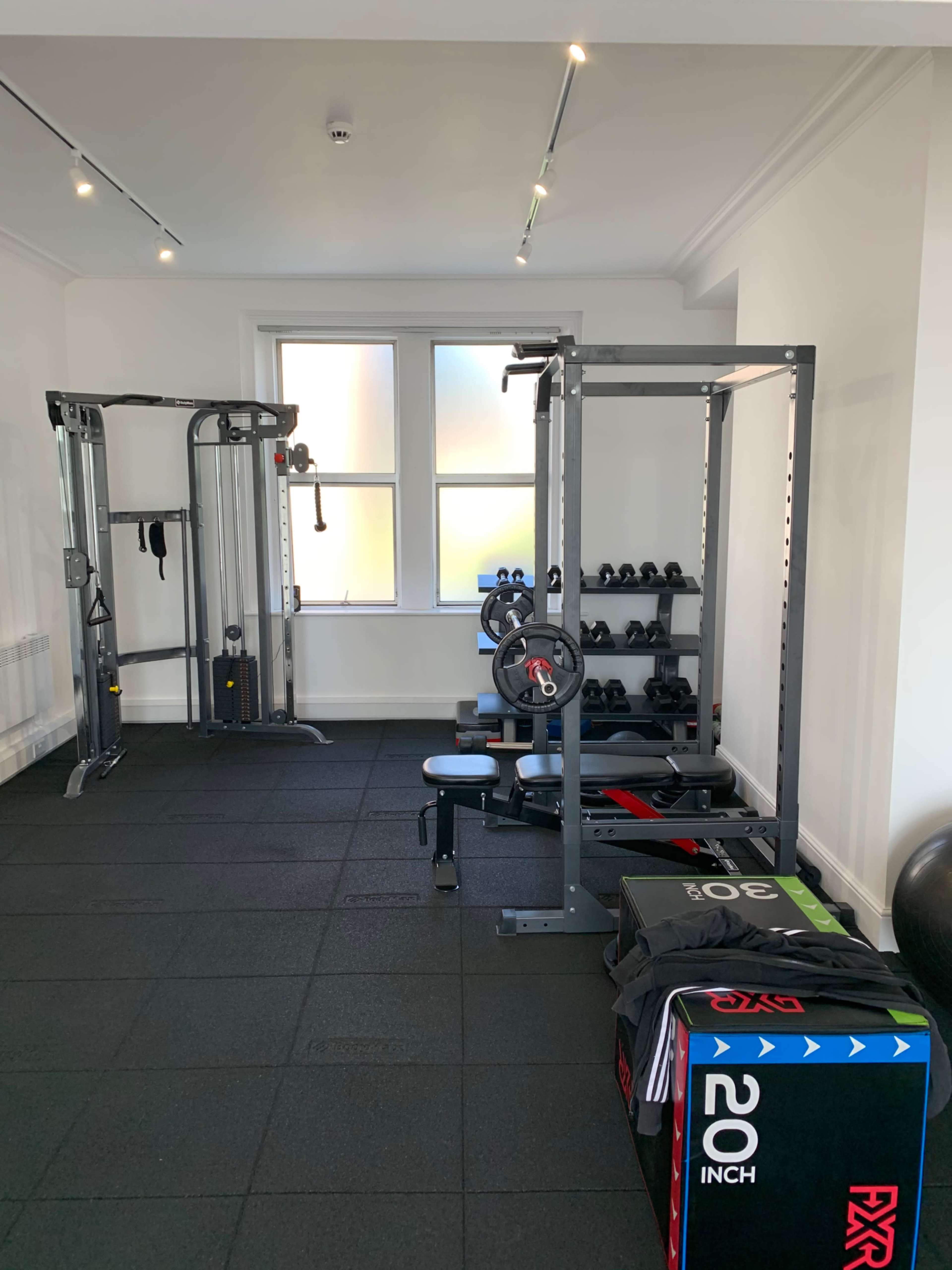 The image shows a gym interior featuring a weight bench, free weights, a multi-gym station, and exercise mats on a dark tiled floor.