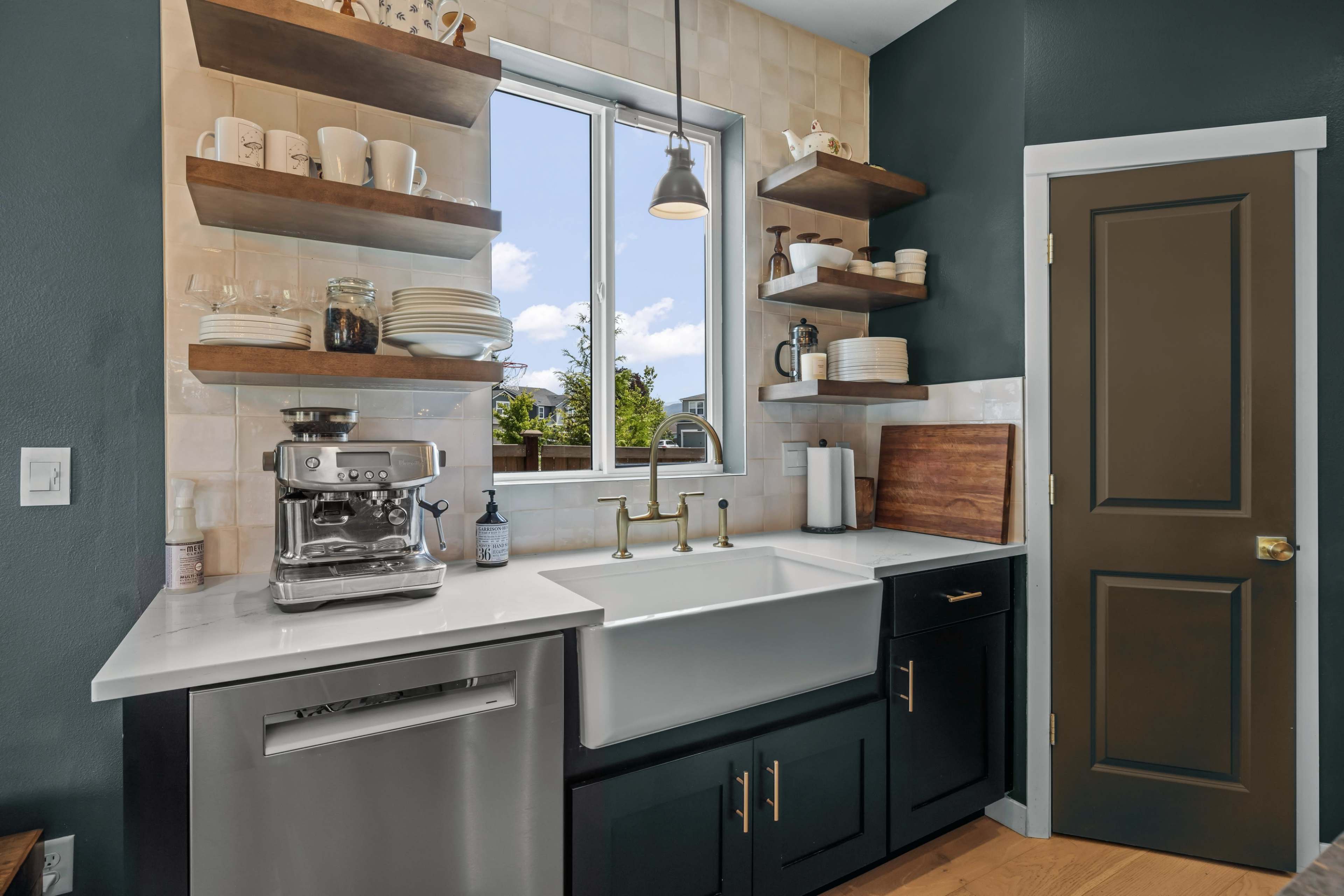 The image shows a modern kitchen corner featuring a farmhouse sink, a coffee machine, open shelf storage with dishes, and a large window.