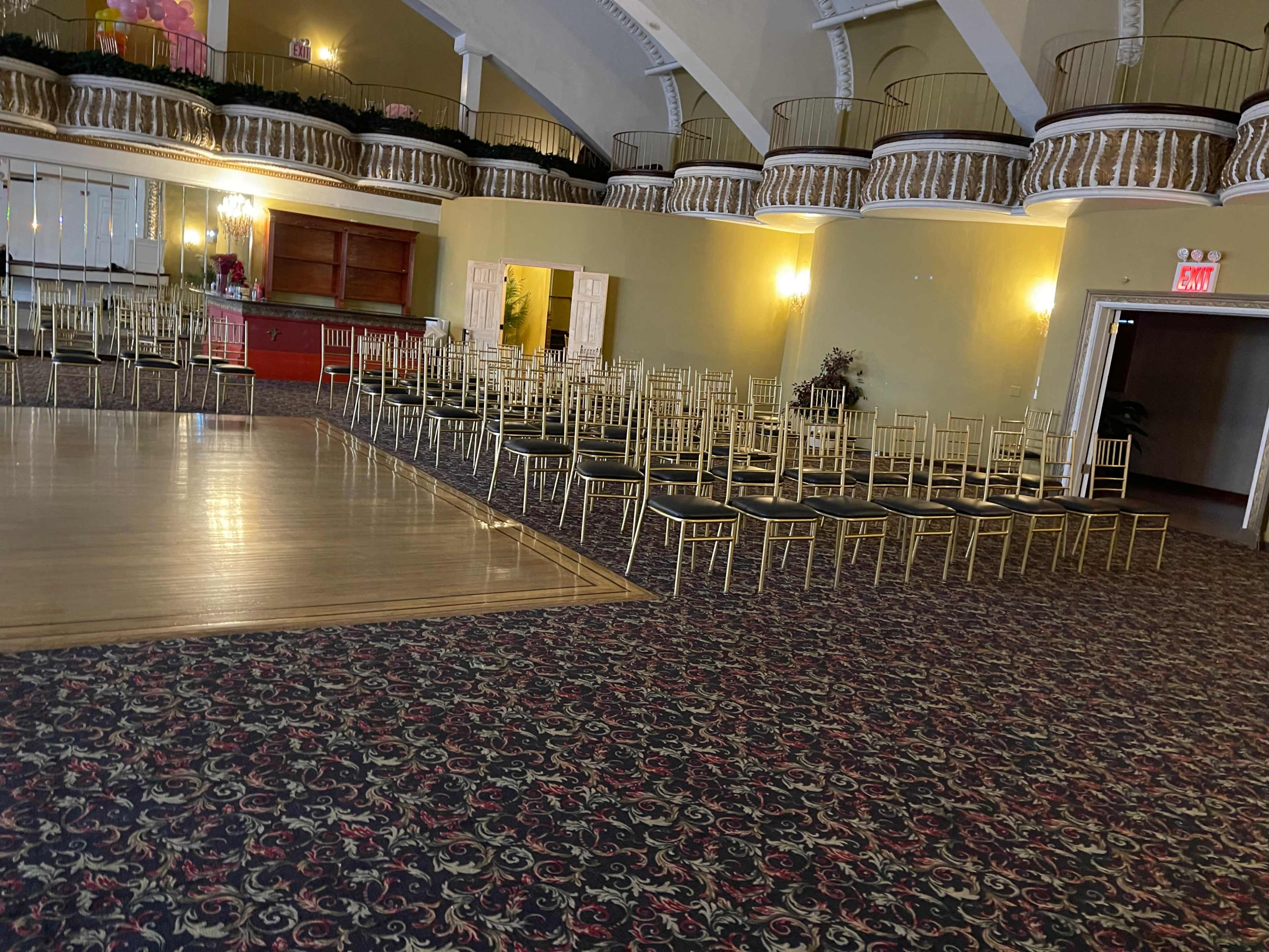 The image shows a large hall with a wooden dance floor, arranged with gold-framed chairs facing a stage area.