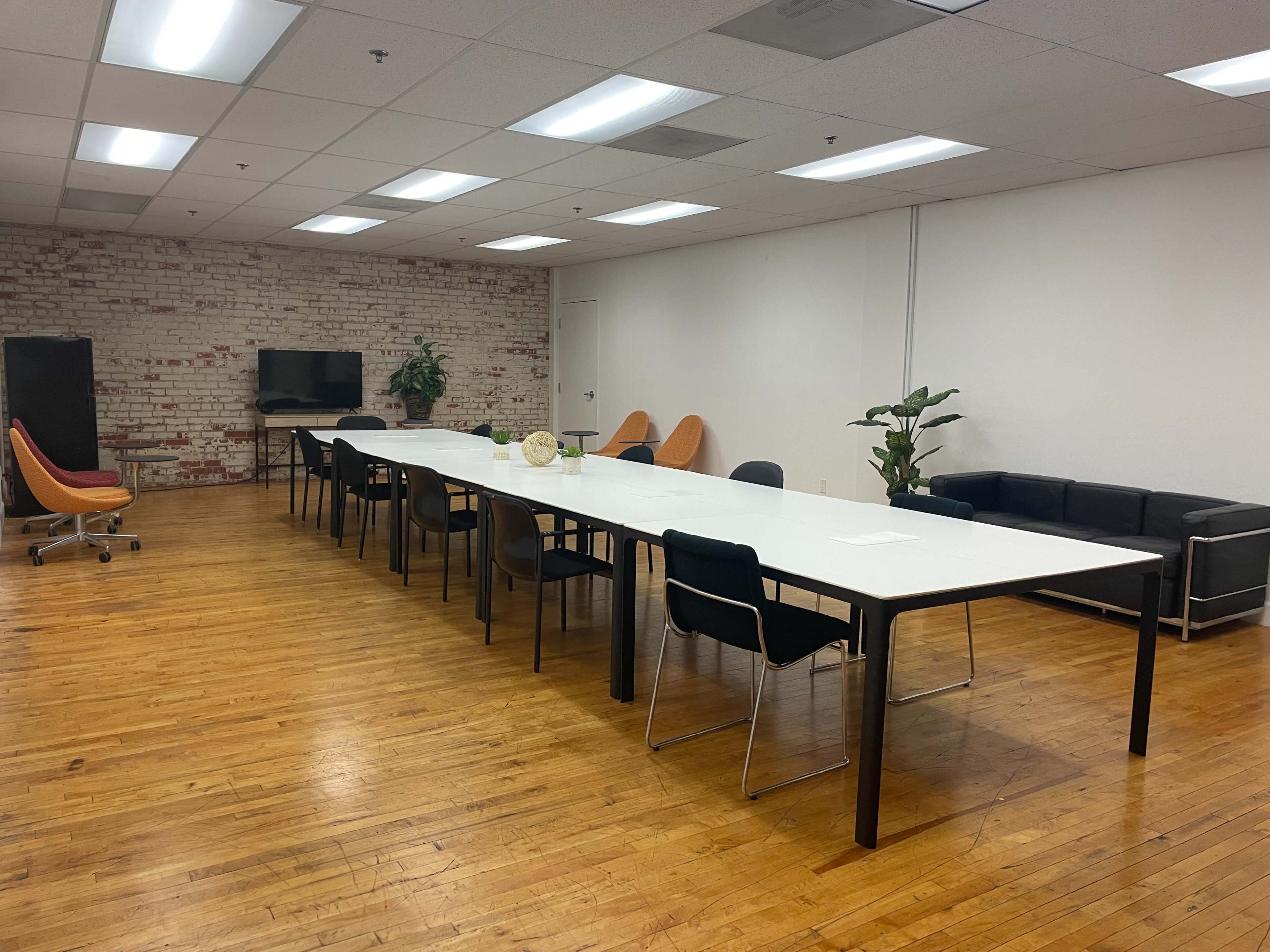 A spacious conference room features a long white table surrounded by chairs, a television on one wall, plants, and seating along the opposite side.