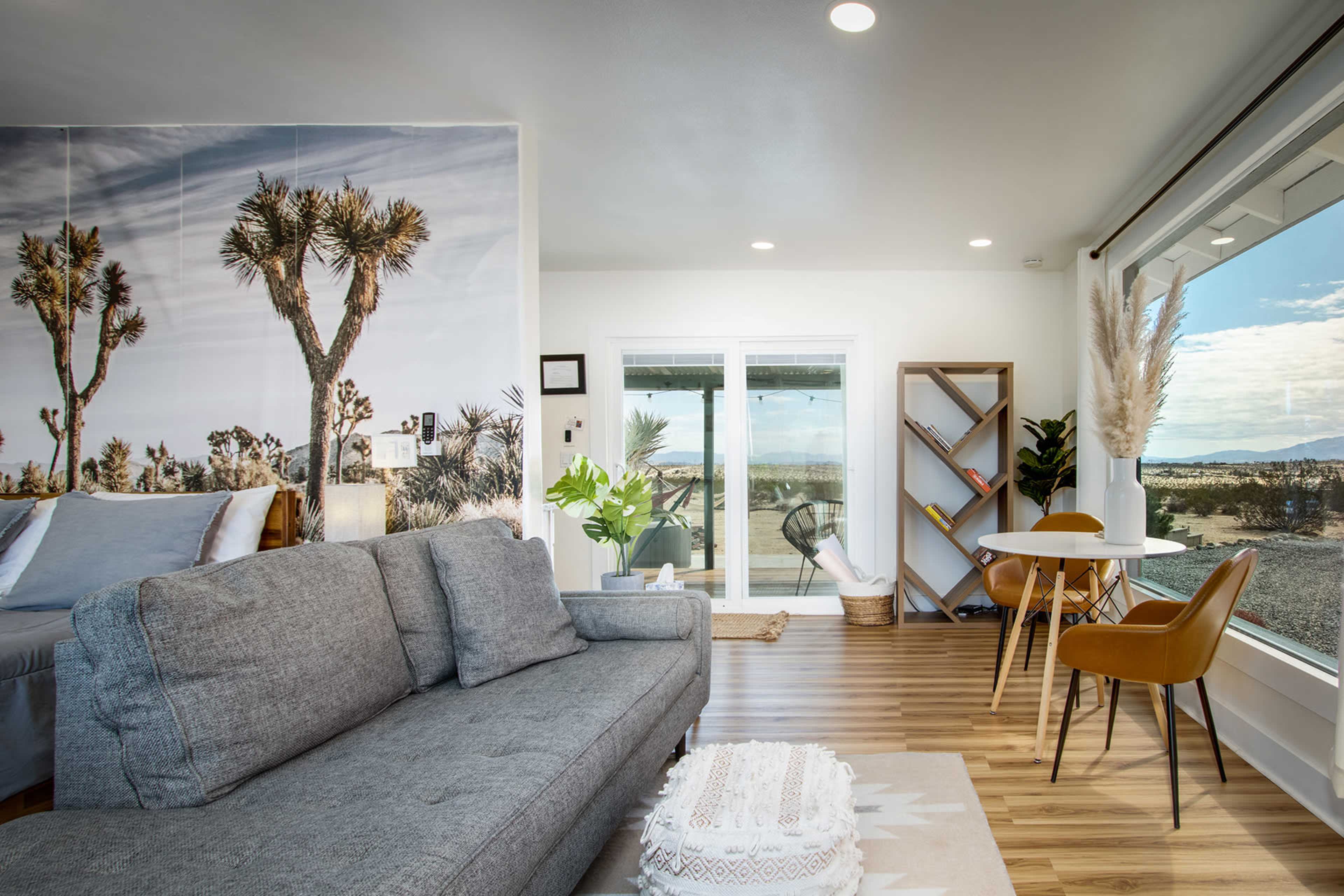 The image shows a modern living space featuring a gray sofa, a dining table with chairs, and large windows that provide a view of a desert landscape.