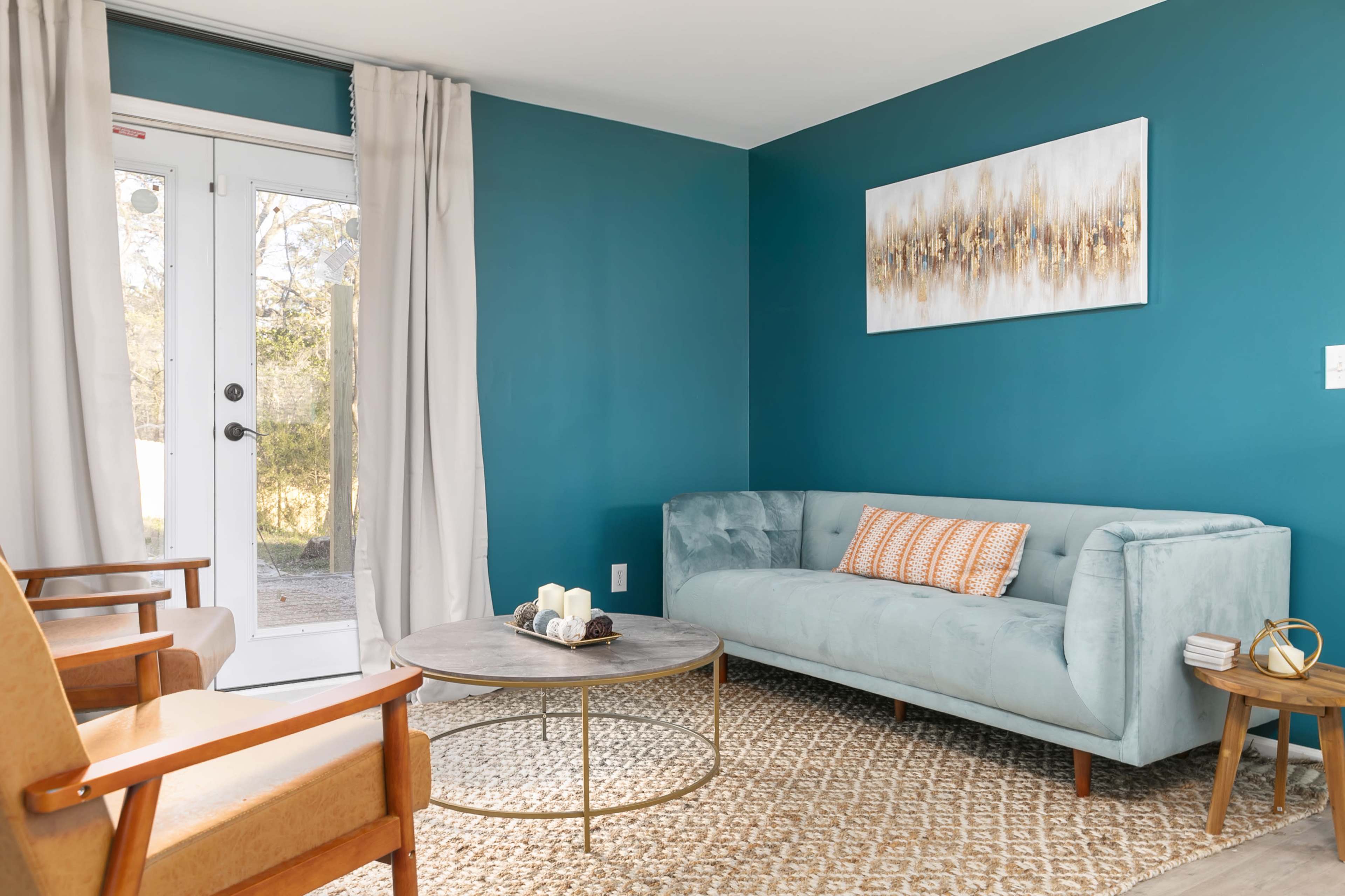 The image shows a minimalist living room with a light blue sofa, a round coffee table, and a patterned rug, near a door leading to an outdoor area.