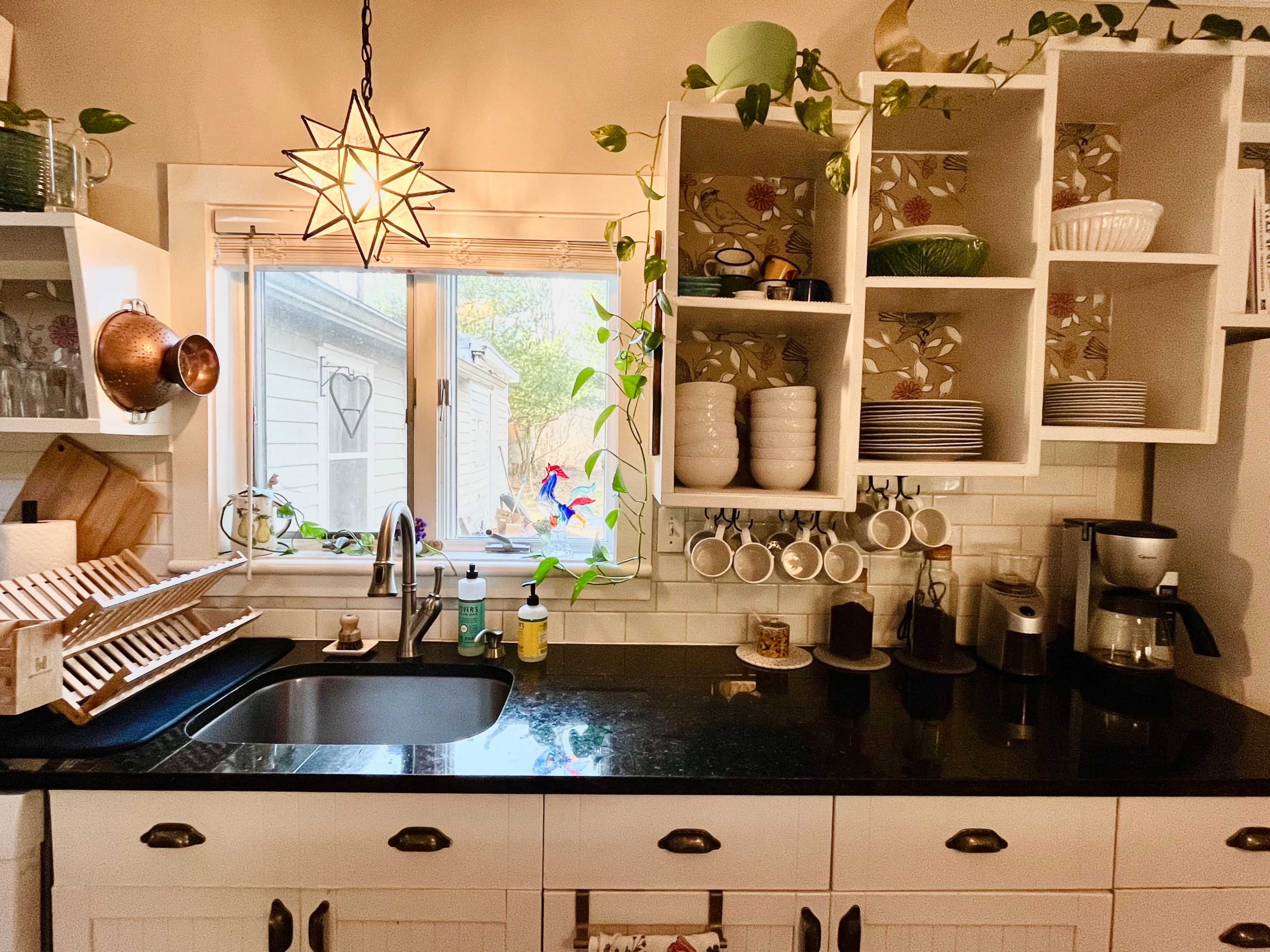 A kitchen countertop with a black sink, drying rack, various dishware, a coffee maker, and a window adorned with plants.