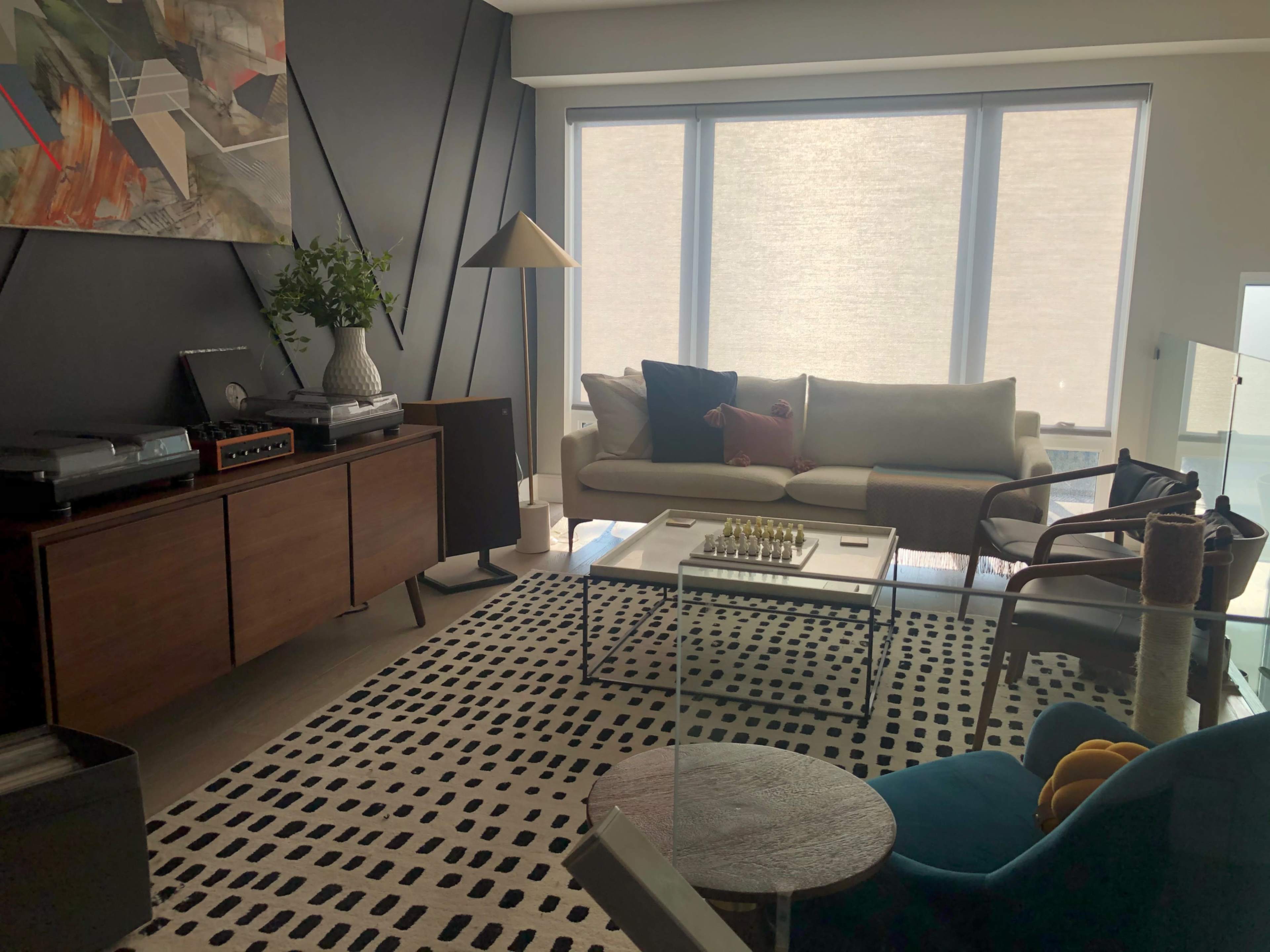 The image depicts a modern living room with a mix of furniture, including a sofa, a coffee table with a chess set, and a patterned rug, all set against a backdrop of large windows and a dark accent wall.
