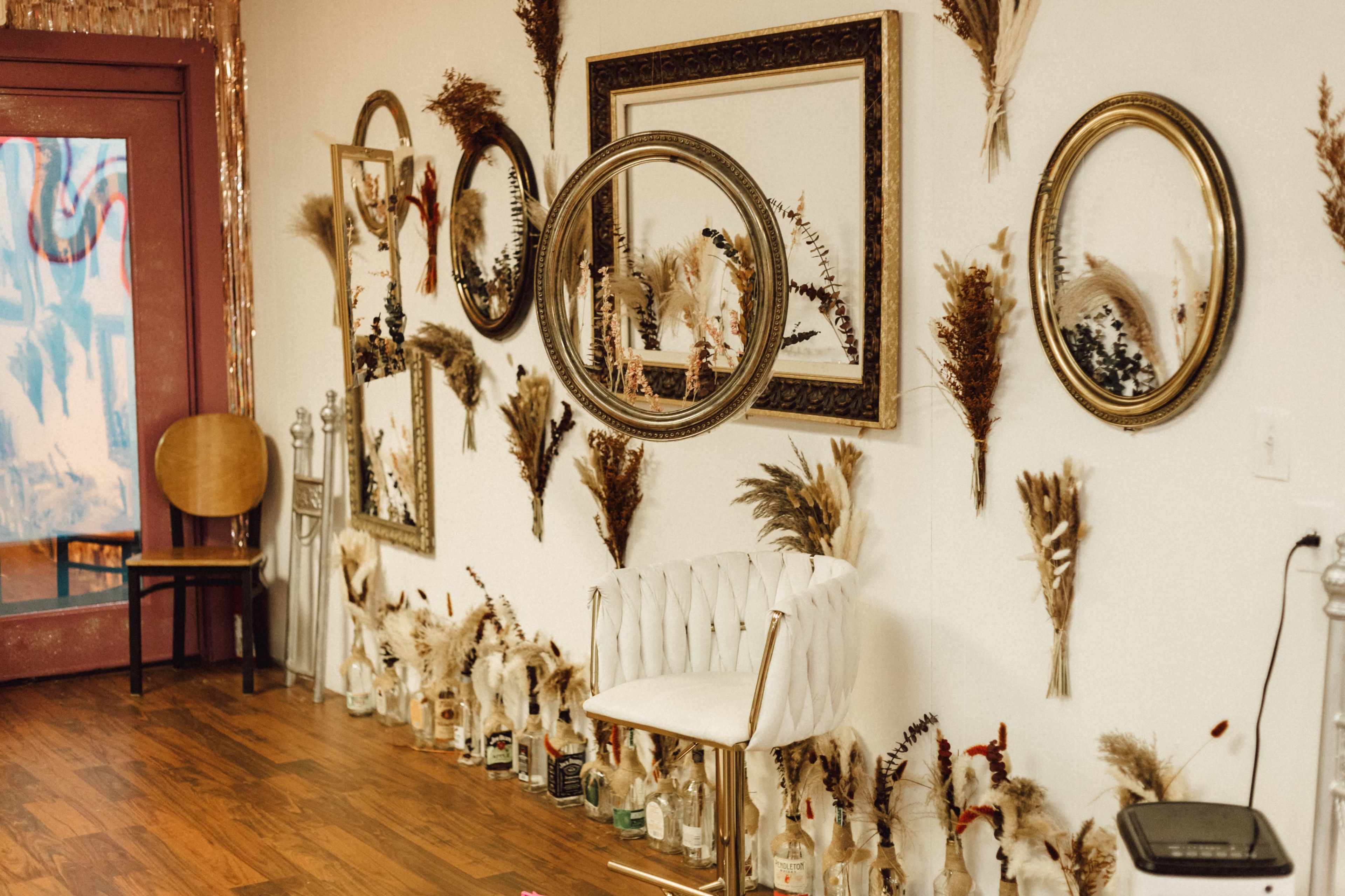 A decorated wall with dried plants arranged around several round mirrors, complemented by a white bar stool in front.