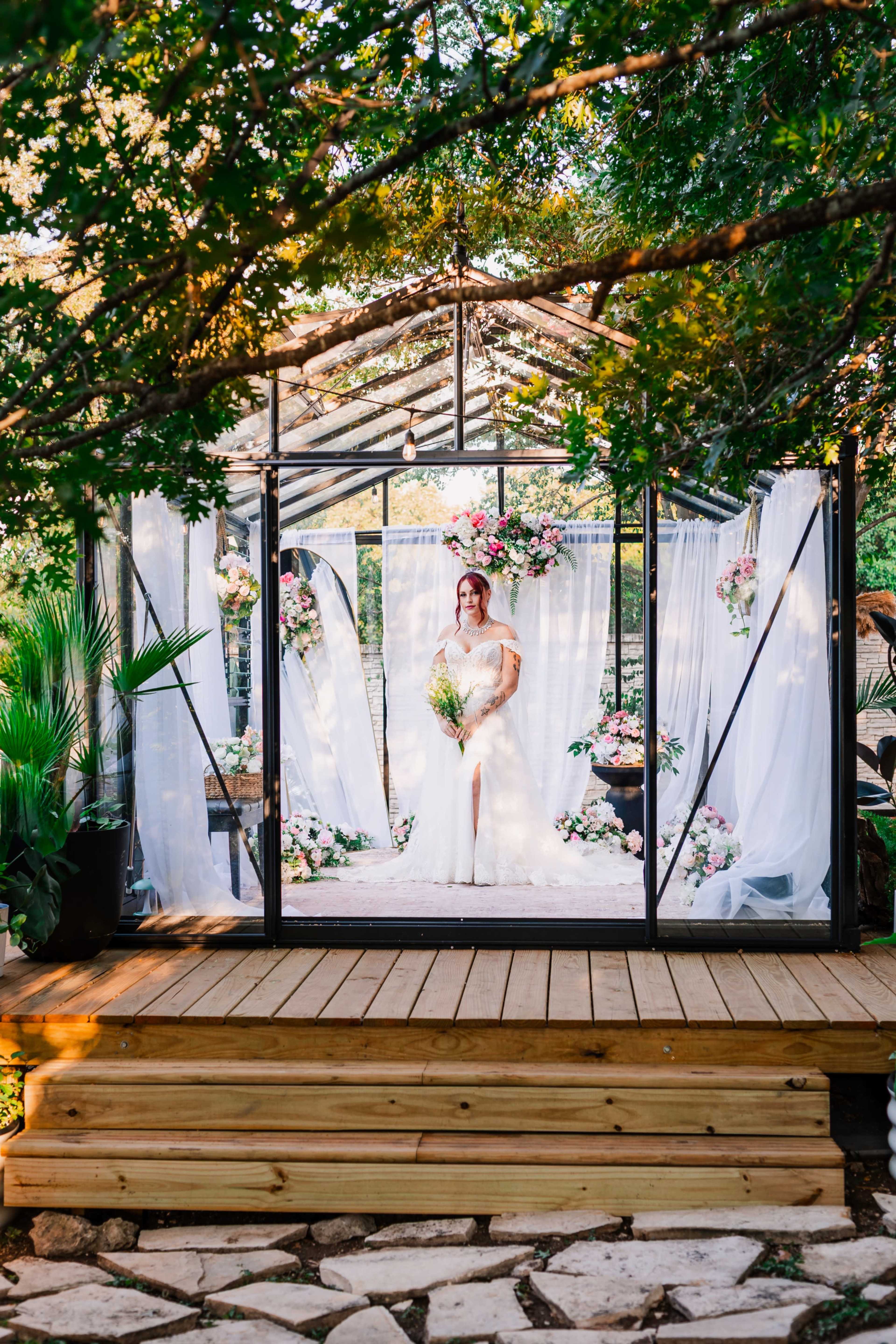 A bride stands in a glass greenhouse adorned with flowers, holding a bouquet, surrounded by greenery.