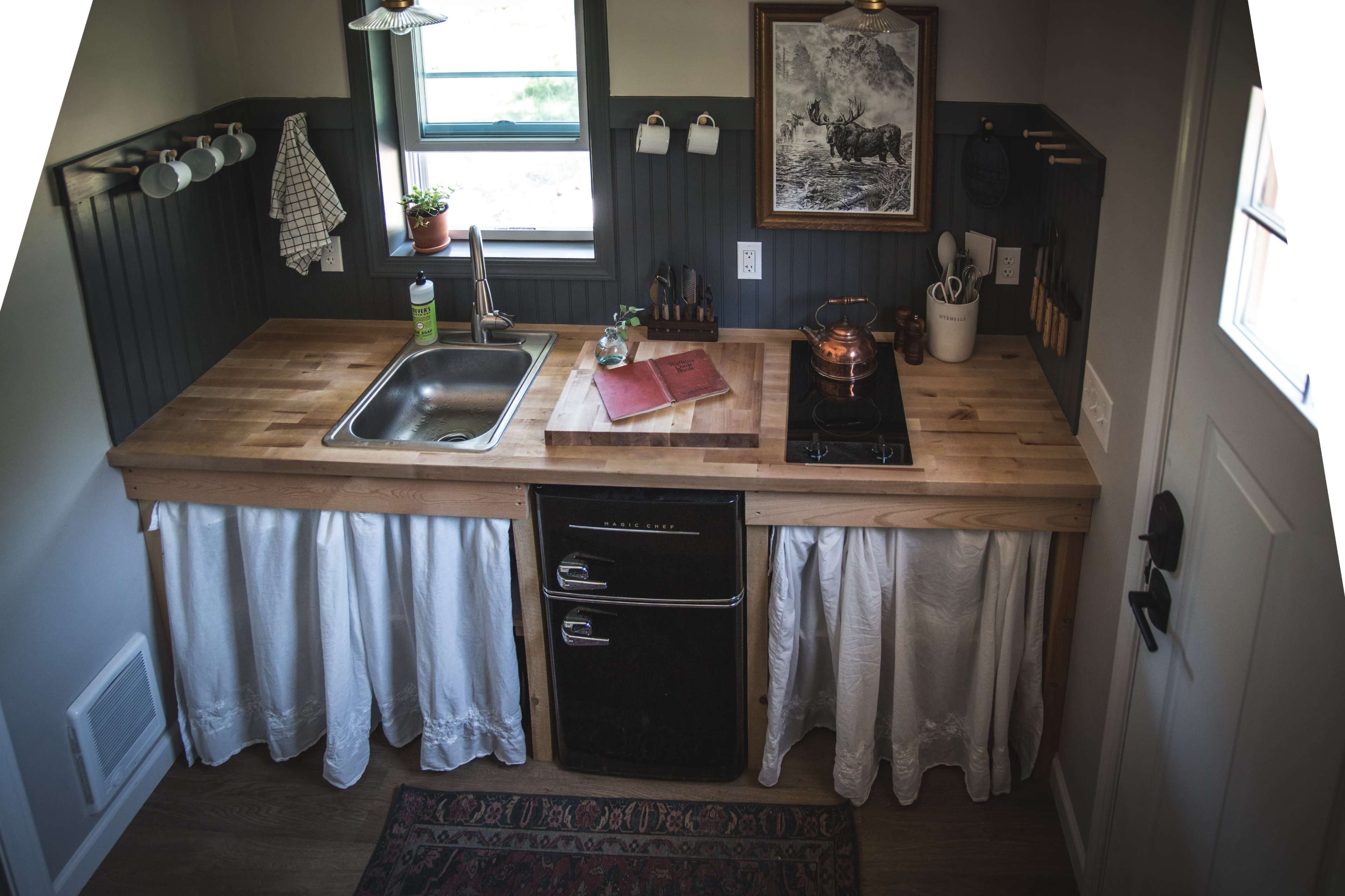 A compact kitchen with a wooden countertop, a sink, a stovetop, and a refrigerator beneath a white curtain, set against dark green wall panels and framed by a door on the right.