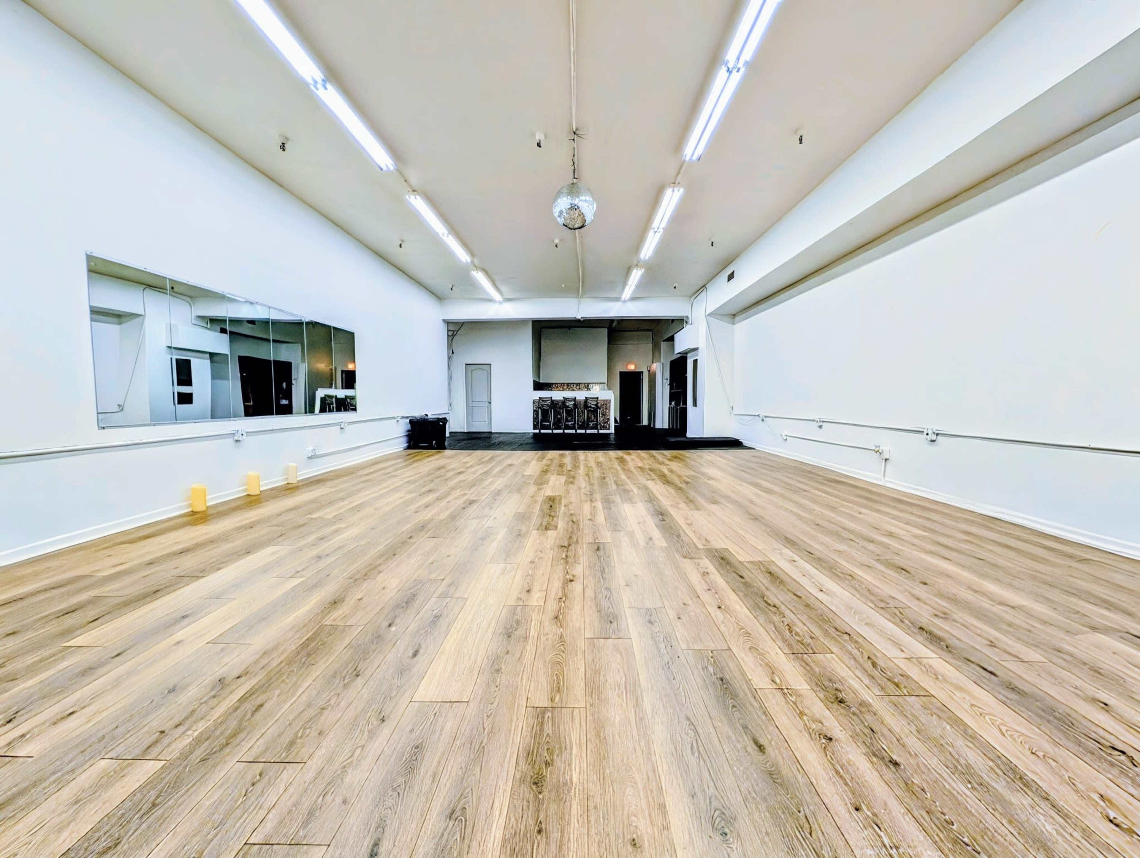 The image shows a spacious, empty dance studio with wooden flooring and mirrors on one wall.