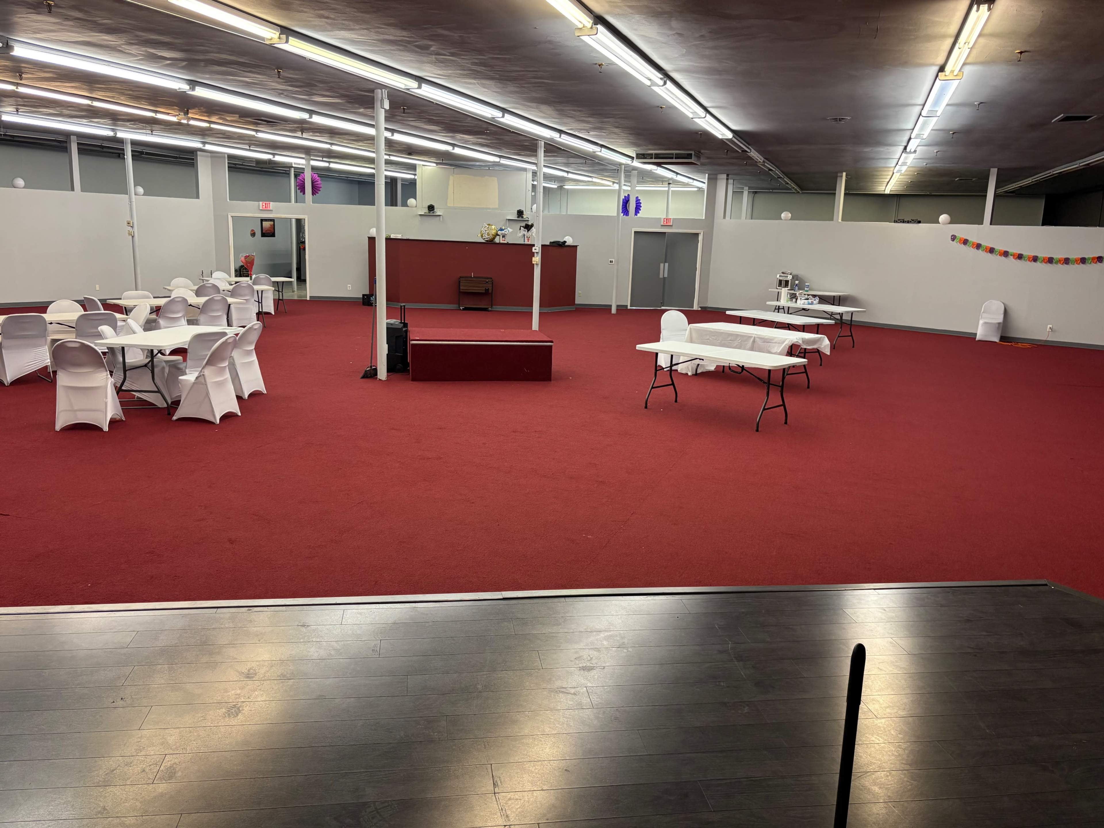 The image shows a spacious, empty event hall with several round tables and chairs arranged around a small elevated stage.