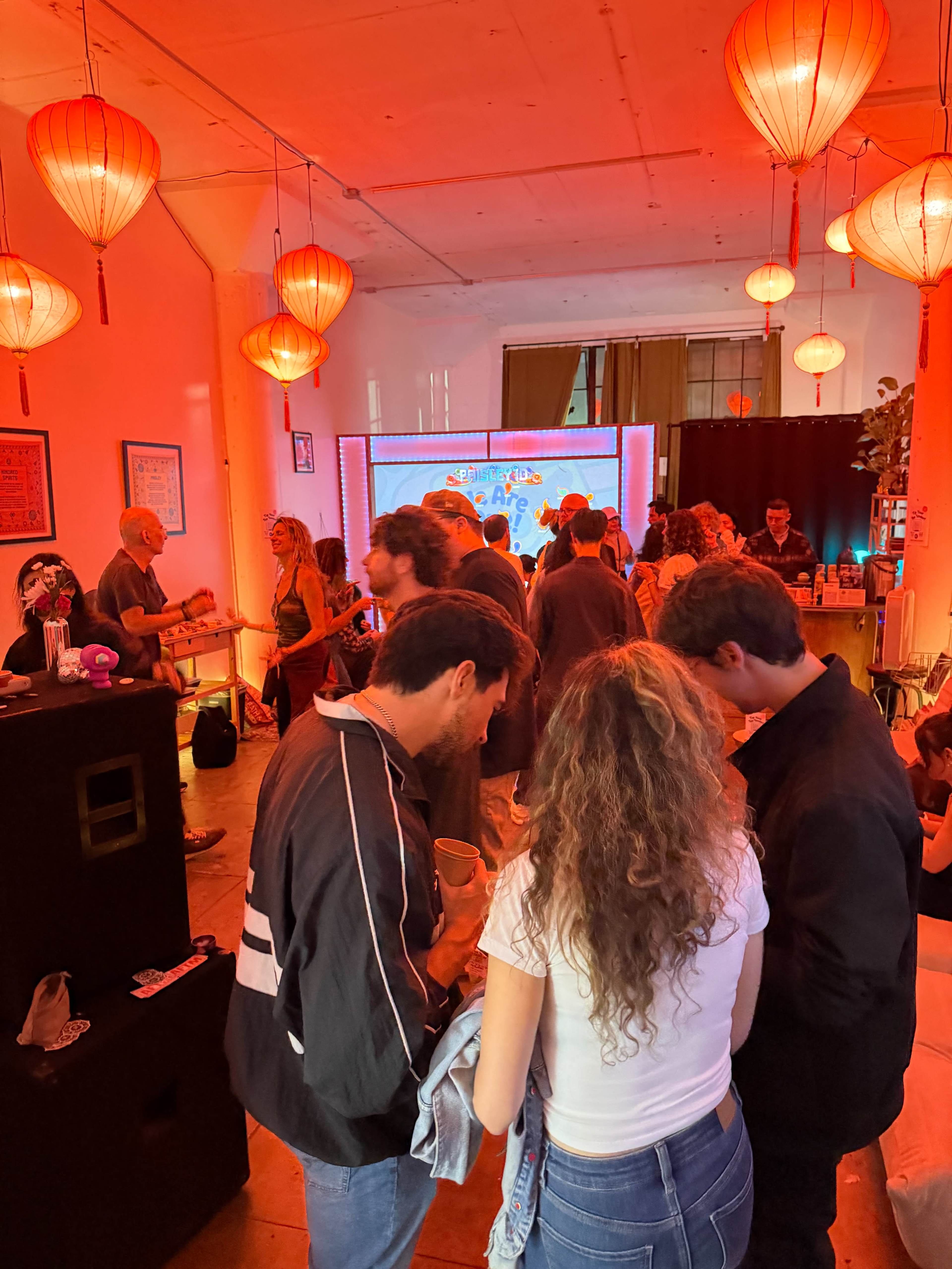 A lively gathering takes place in a warmly lit room adorned with red lanterns, featuring a projector screen and a crowd of people mingling.