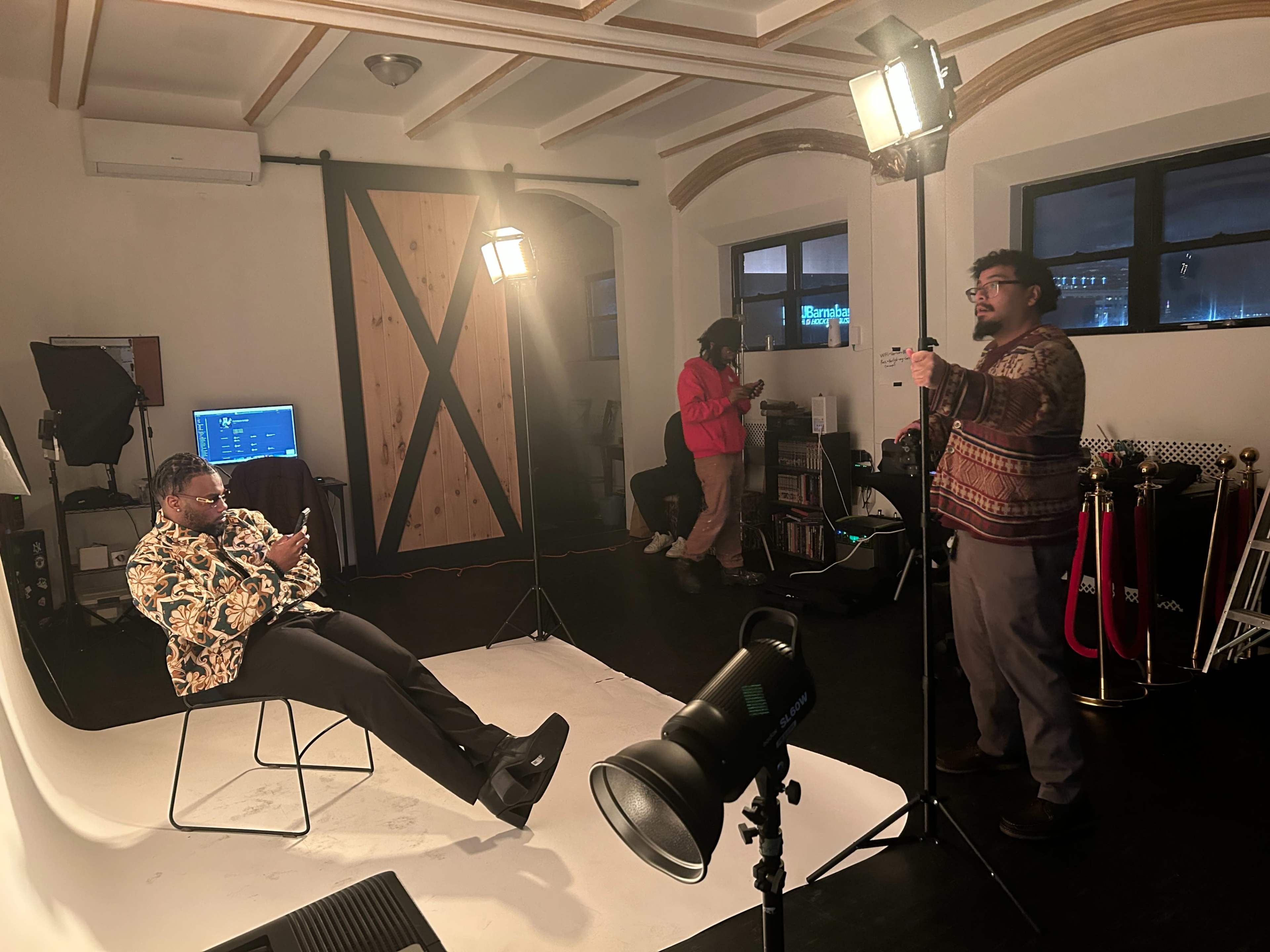 A photoshoot setup features a person sitting on a chair while another appears to be adjusting lighting equipment in a studio with a backdrop.