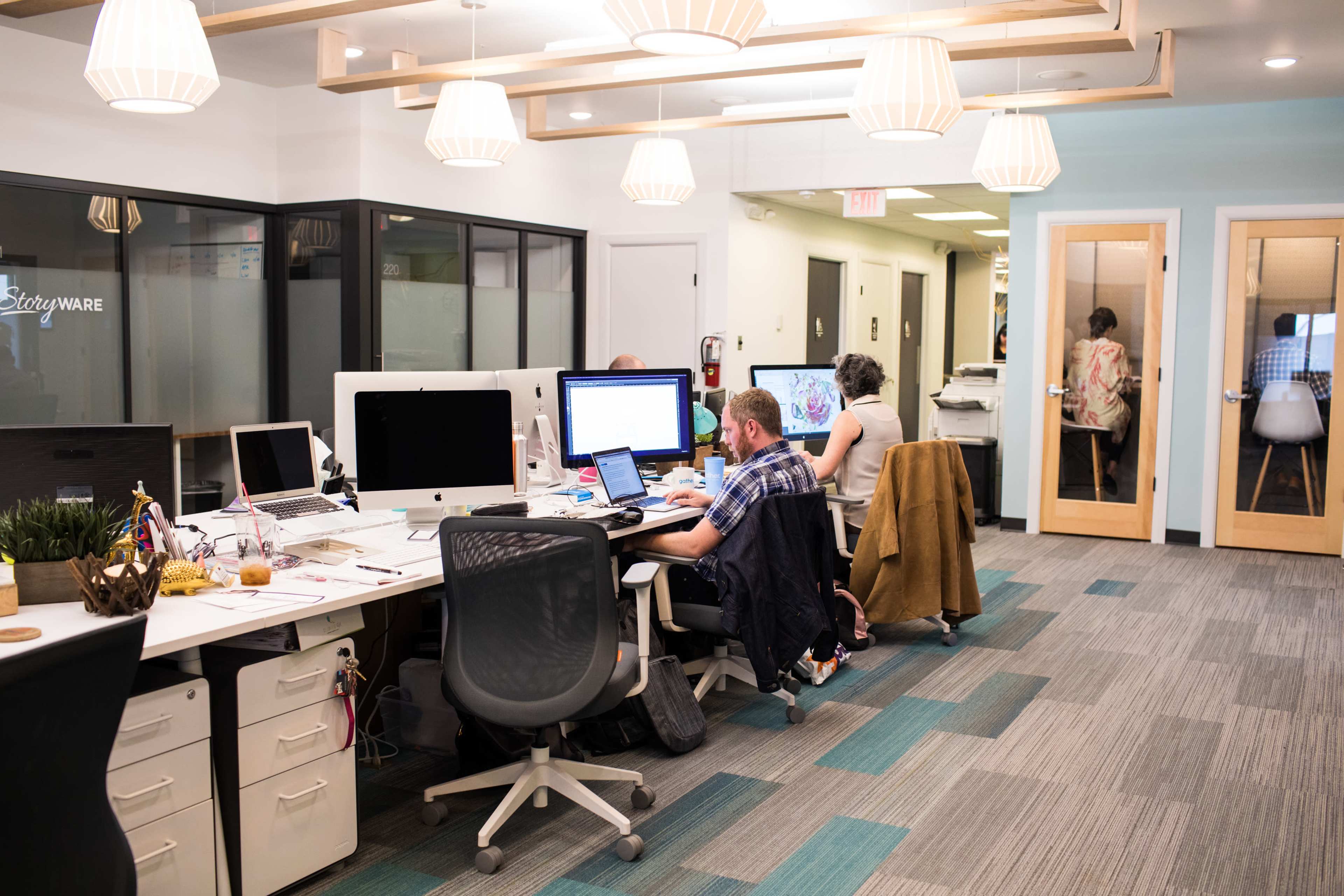 A modern office space with several workstations, each equipped with desktop computers, and two individuals working at their desks while a third person is seen in a meeting room.