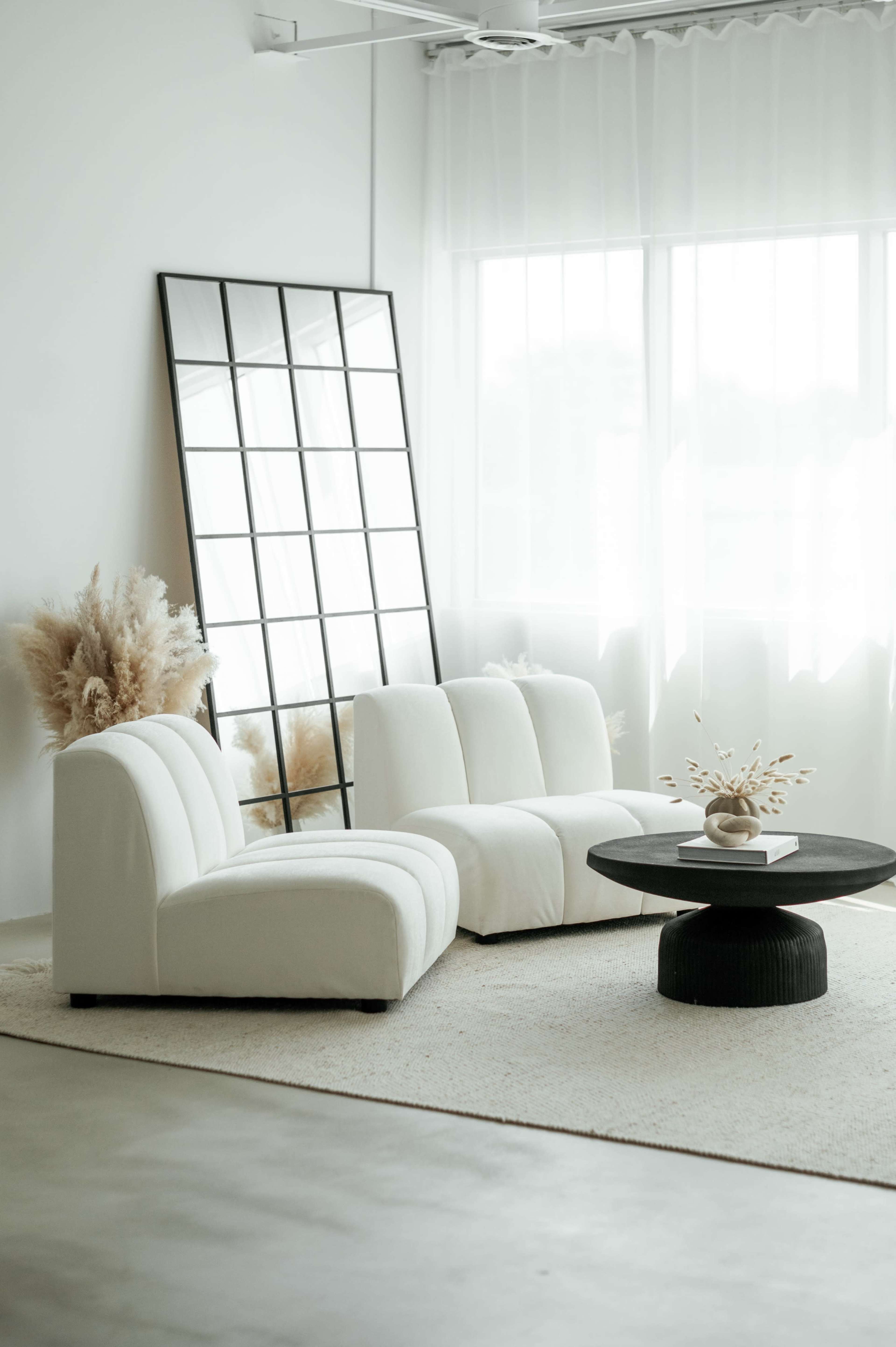 A minimalist living room with white upholstered sofas, a round black coffee table, and a large mirror reflecting natural light.