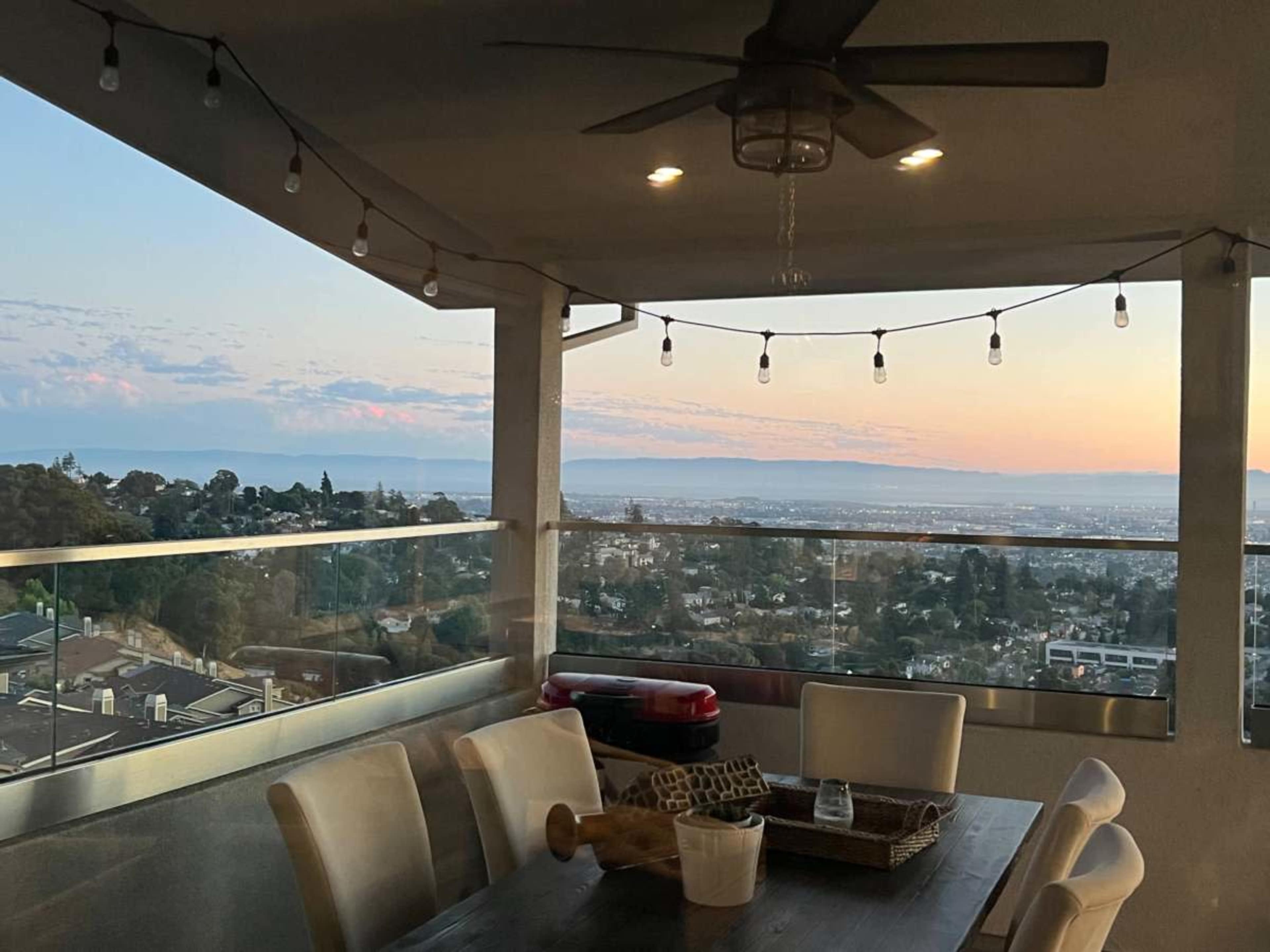 A dining area on a balcony with a clear view of the landscape and sunset, featuring a table set for meals and string lights overhead.
