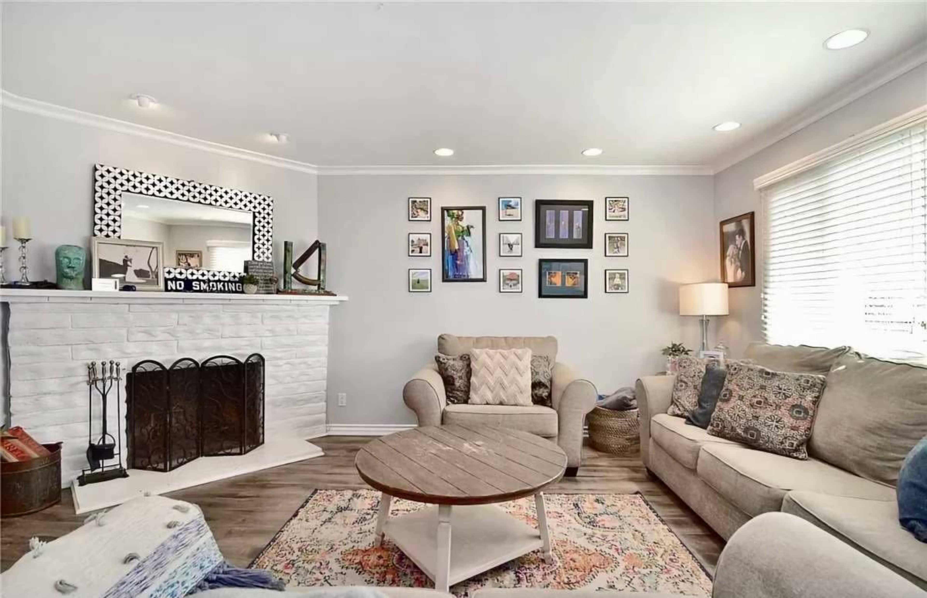 The image shows a cozy living room with a light-colored couch, a round wooden coffee table, a white brick fireplace, and various framed pictures on the walls.