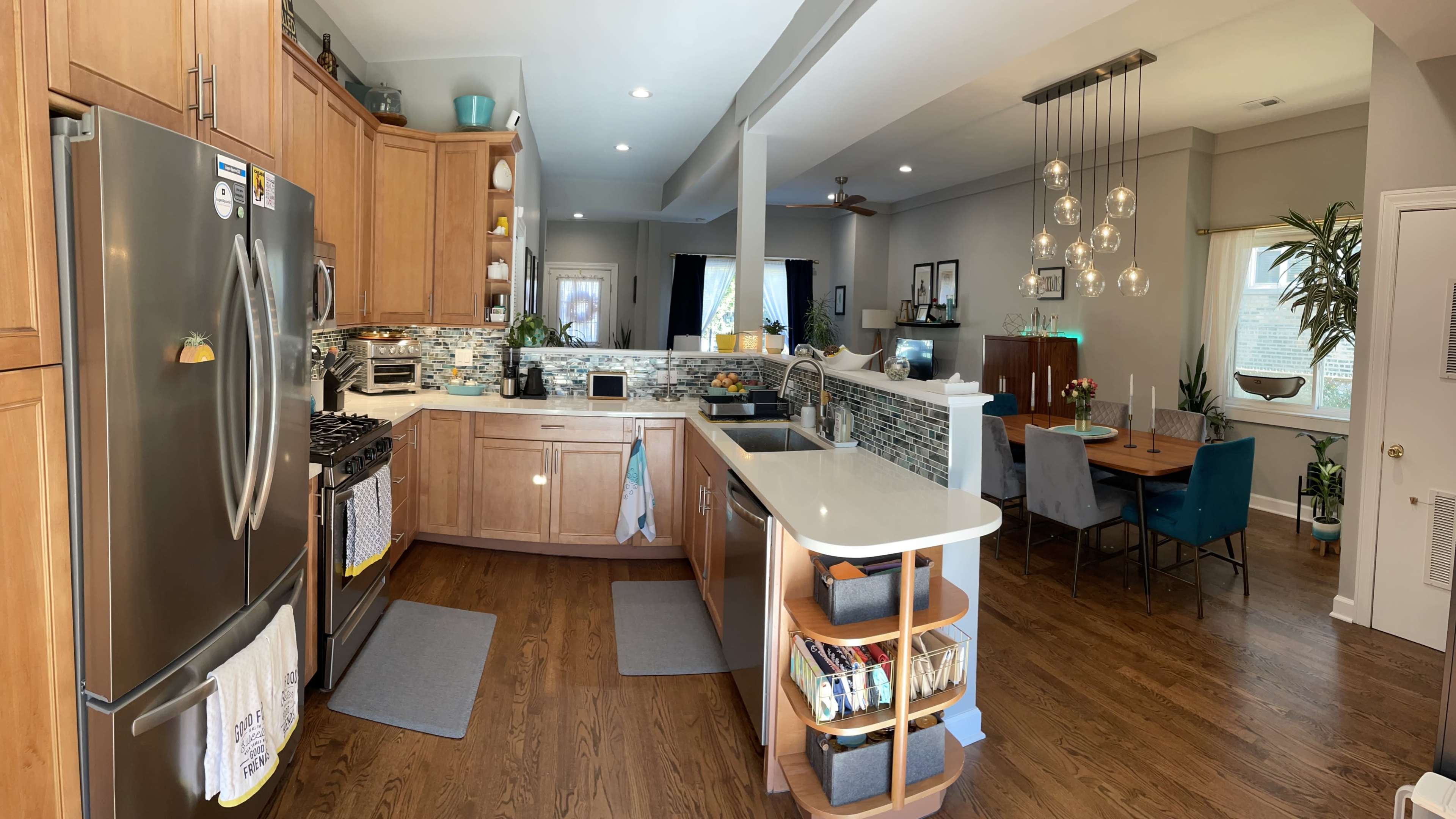 A modern kitchen with wooden cabinetry, stainless steel appliances, and an island with seating, adjacent to a dining area featuring a table and hanging light fixtures.