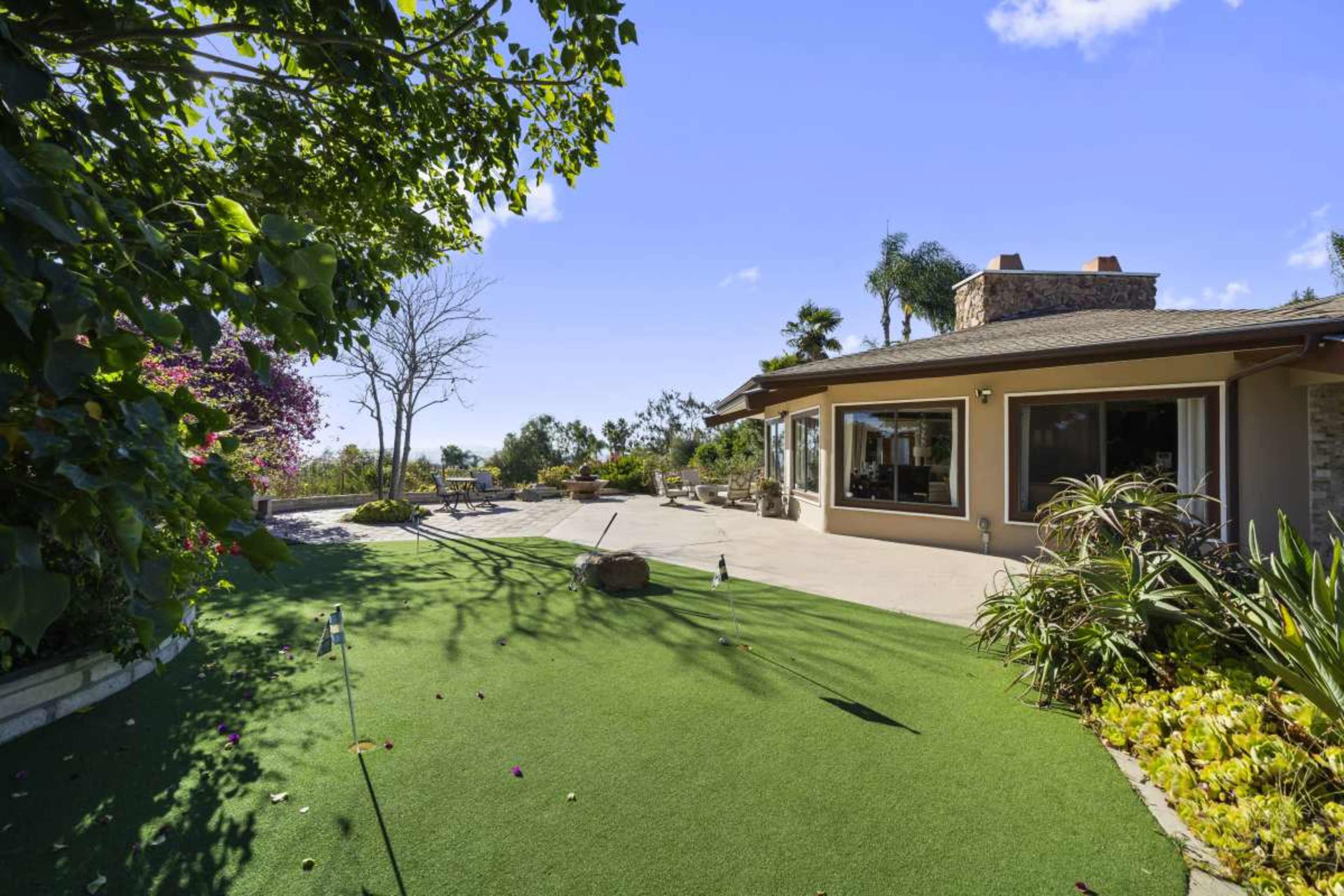 The image shows a landscaped backyard with a putting green, surrounded by trees and shrubs, and a house with large windows and a stone chimney.