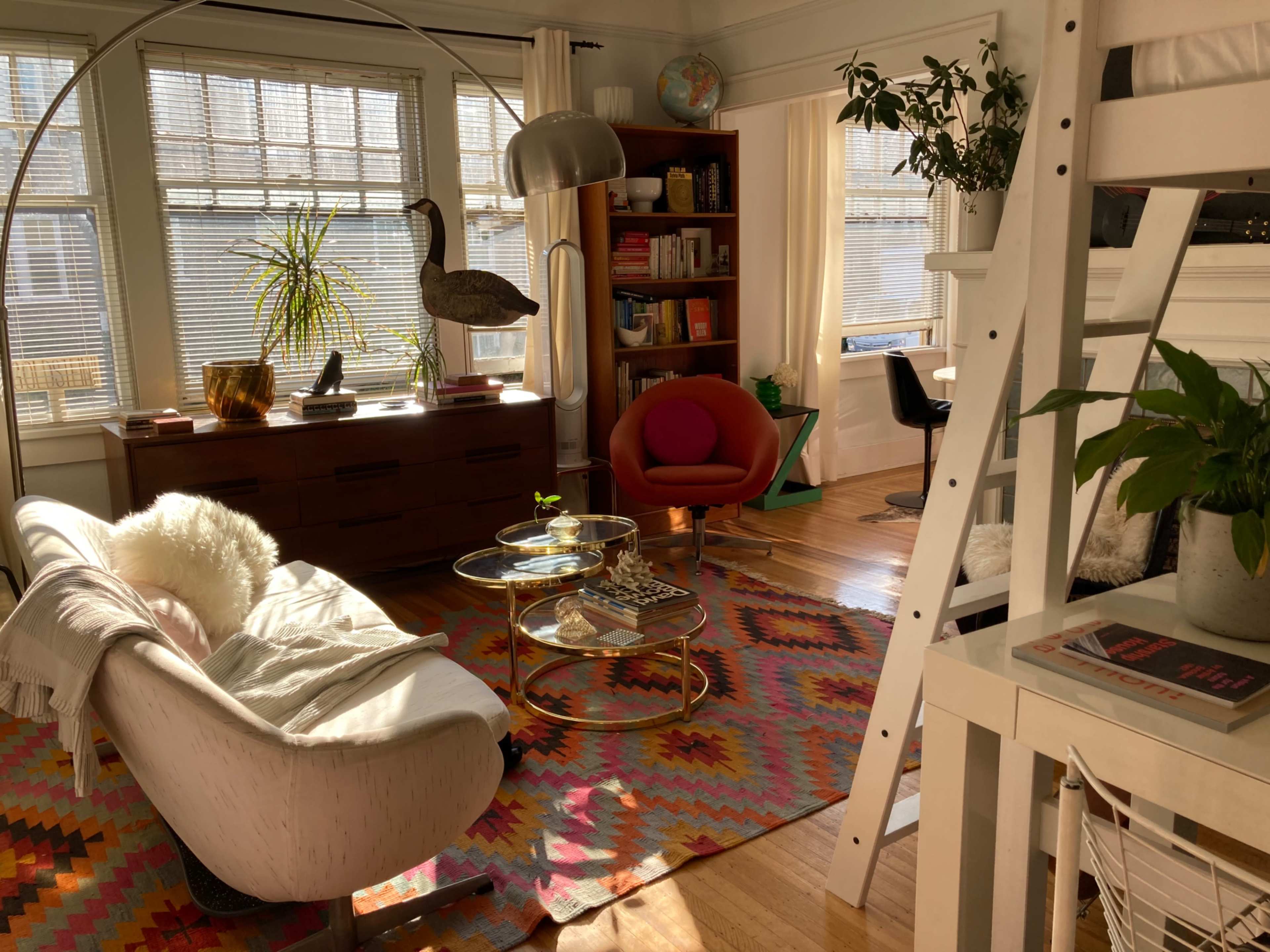 A bright living room with a curved sofa, a round coffee table, a red accent chair, and a bookshelf, illuminated by natural light through large windows.