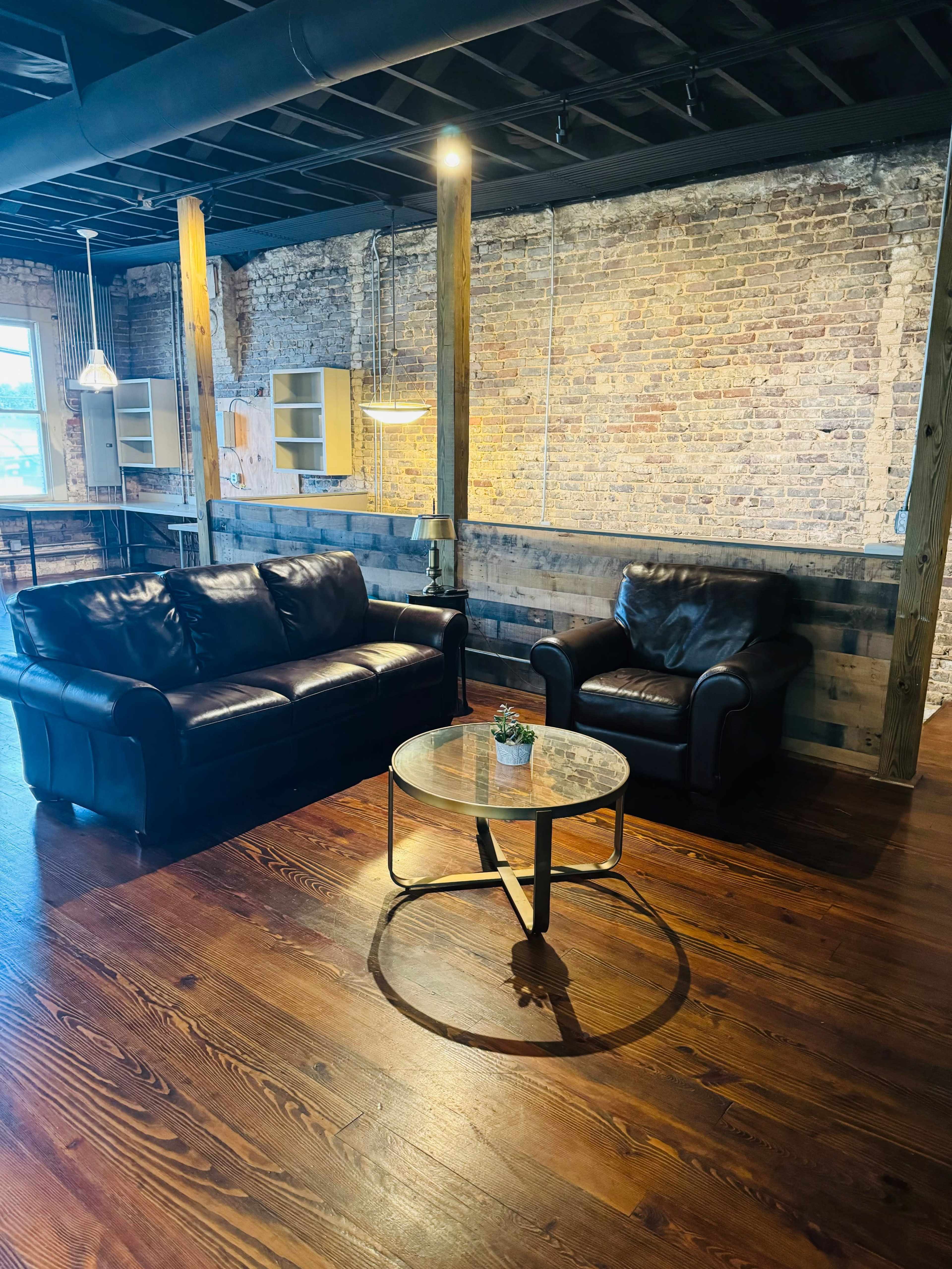 A modern seating area with a black leather sofa and armchair positioned around a circular glass coffee table, set against a backdrop of exposed brick walls and wooden beams.