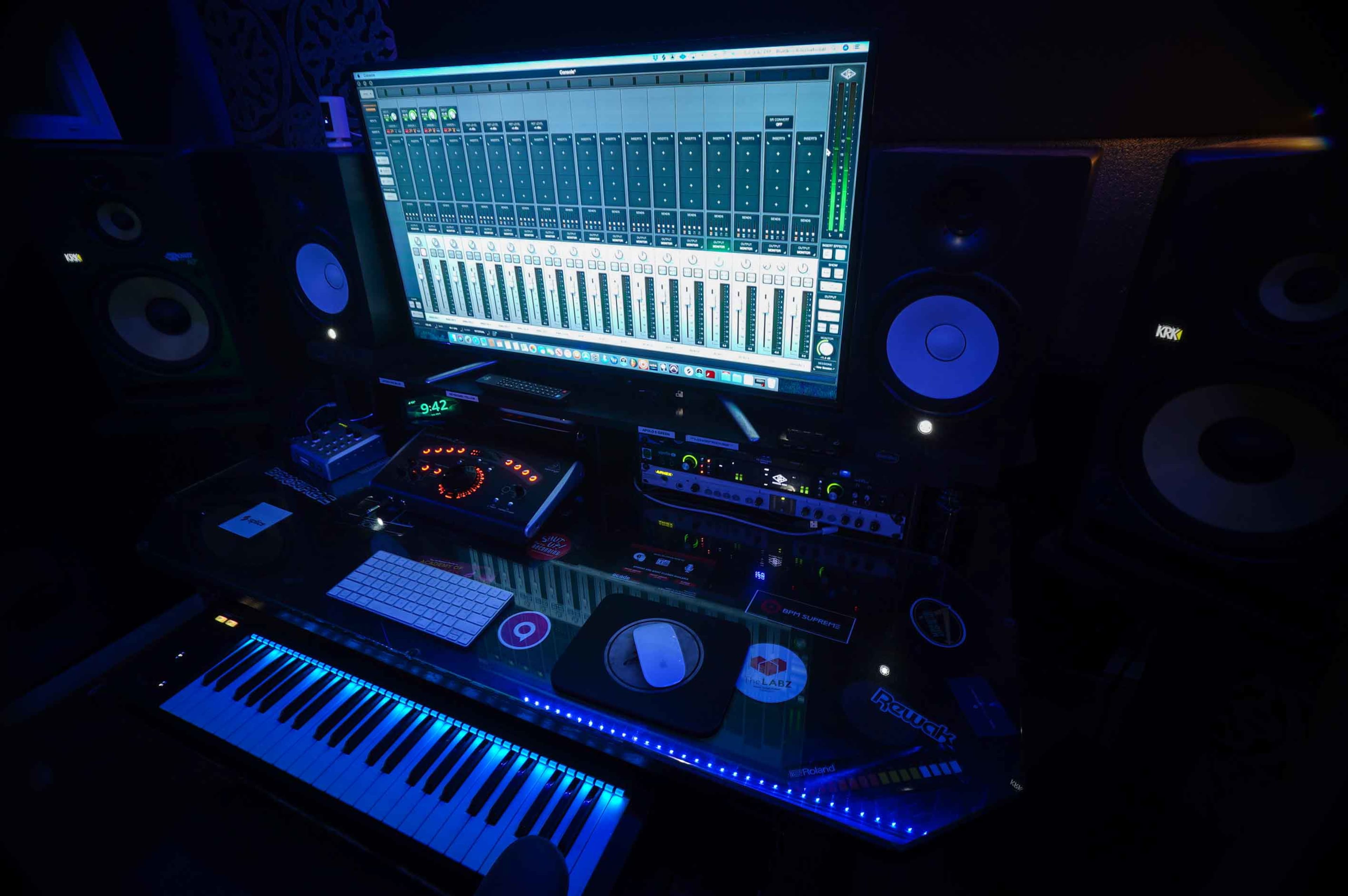 A music production workstation features a keyboard, audio interface, and a large computer monitor displaying a digital audio mixer, all set against a backdrop of low lighting.