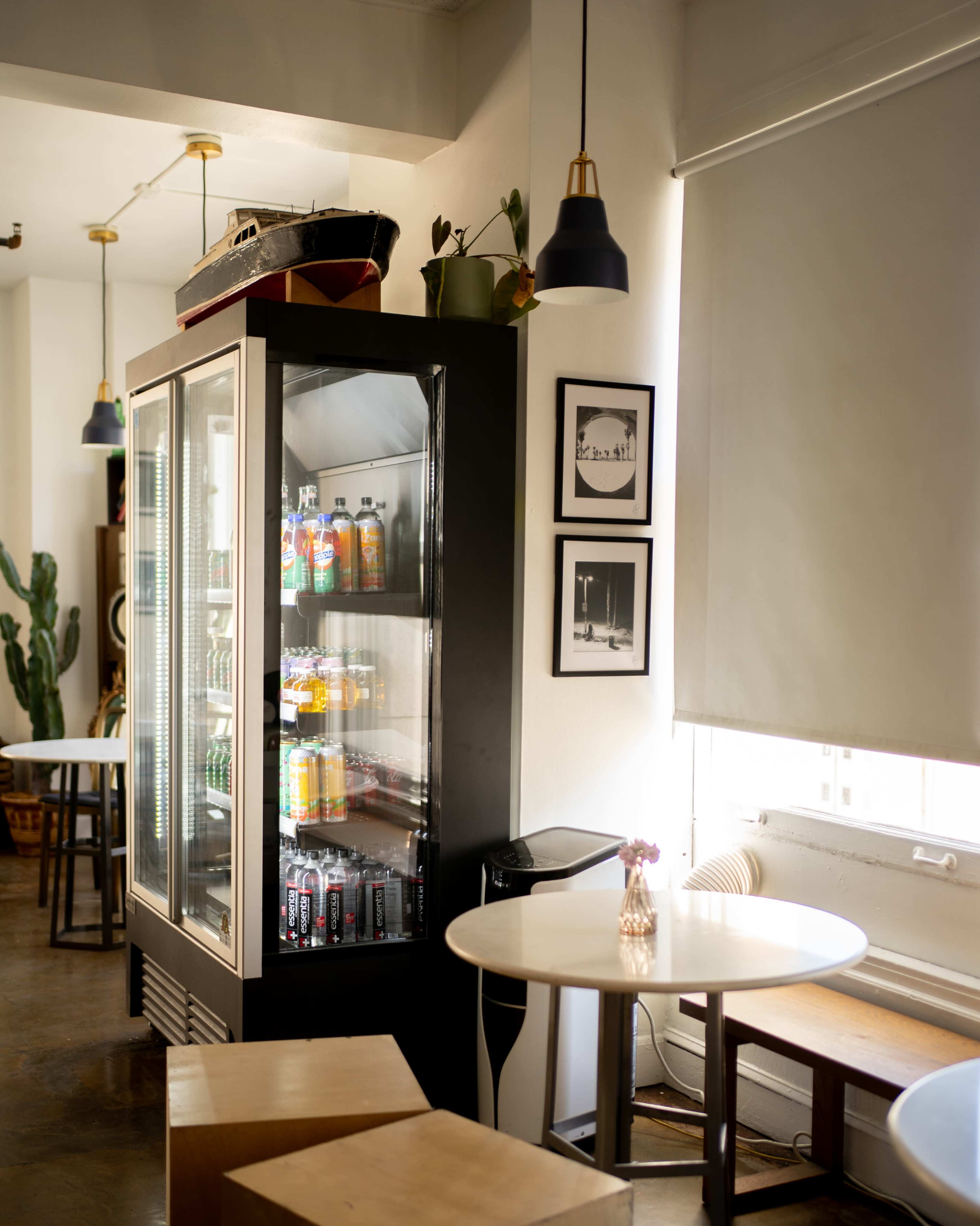The image shows a modern café interior with a glass display refrigerator filled with drinks, surrounded by wooden tables and chairs.